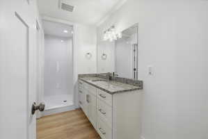 Bathroom featuring a walk-in shower with white surround, a vanity with a speckled countertop and integrated sink, a rectangular mirror, and wood-finish flooring