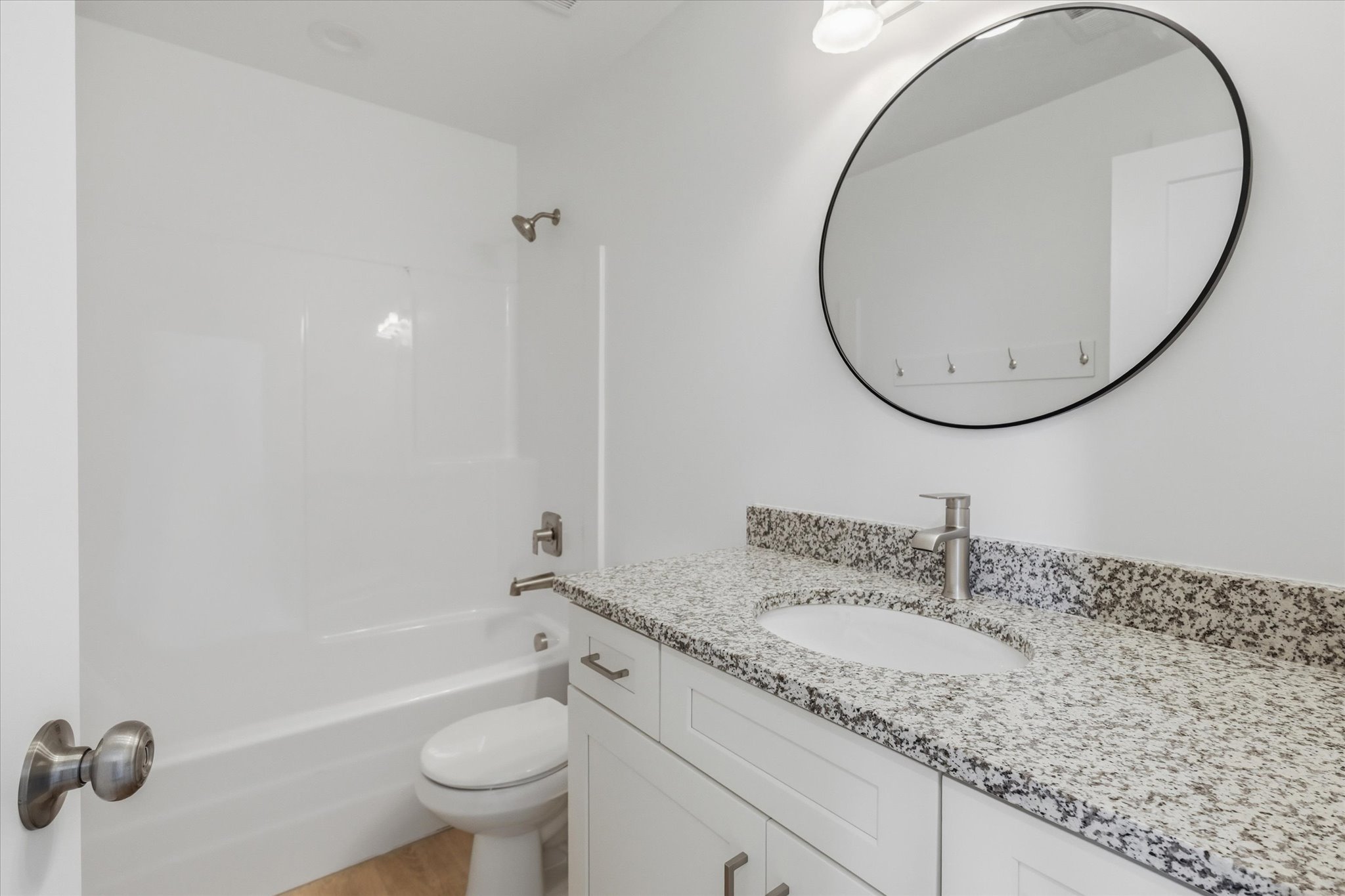 Bright bathroom featuring a white vanity with shaker-style cabinetry, a speckled countertop, an undermount sink with a brushed nickel faucet, a large oval mirror with a black frame, and a white bathtub with a built-in shower