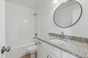 Bright bathroom featuring a white vanity with shaker-style cabinetry, a speckled countertop, an undermount sink with a brushed nickel faucet, a large oval mirror with a black frame, and a white bathtub with a built-in shower