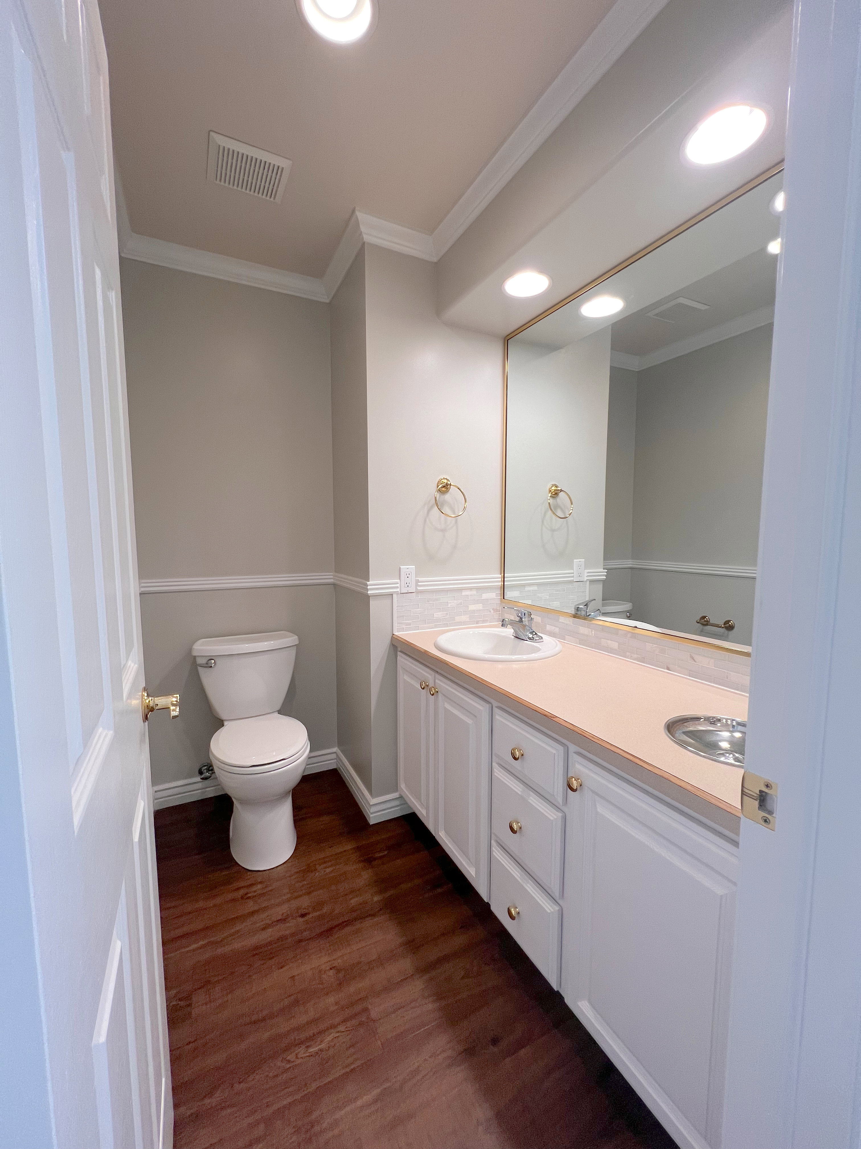 Bathroom with wood-finish flooring, a white vanity featuring a single sink and brass-toned hardware, a full-width wall mirror, and recessed ceiling lighting