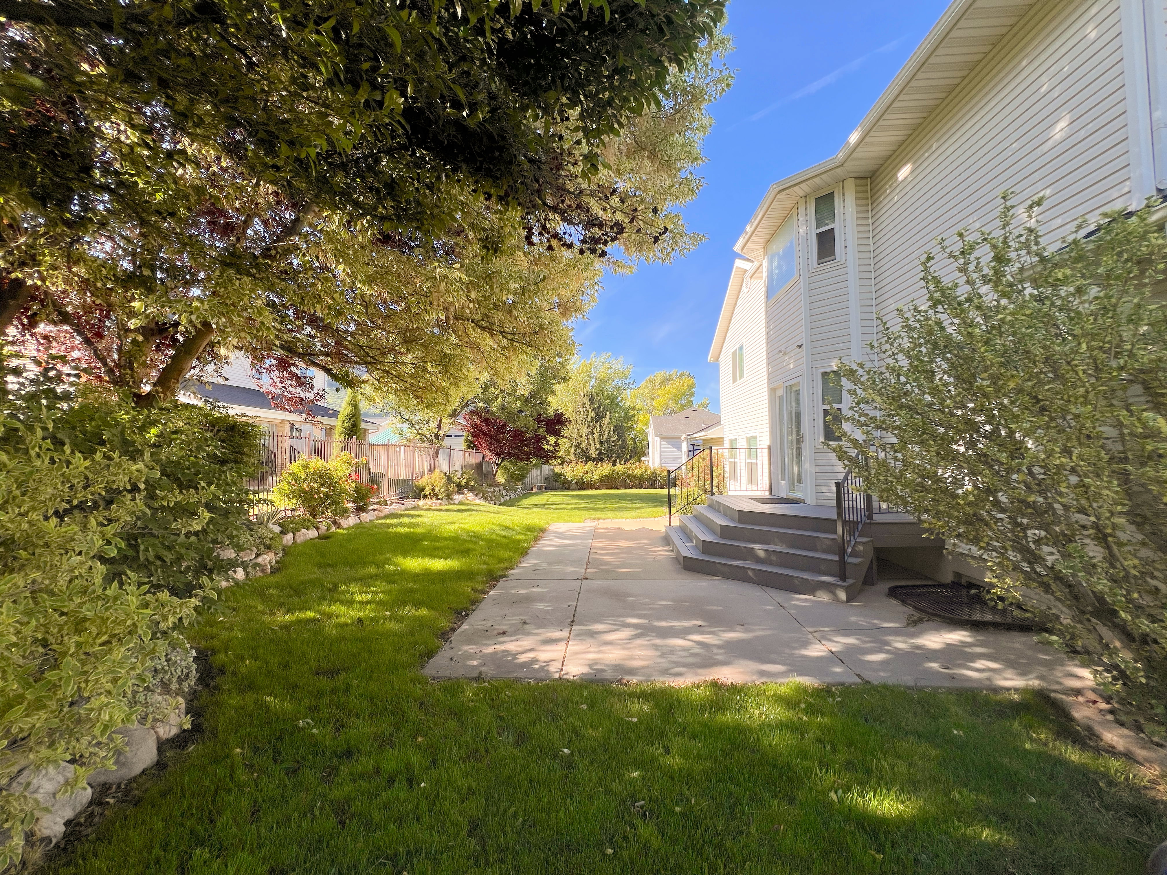Rear exterior featuring a concrete patio, multi-tiered steps with black metal railings, and a expansive lawn with mature trees and shrubbery
