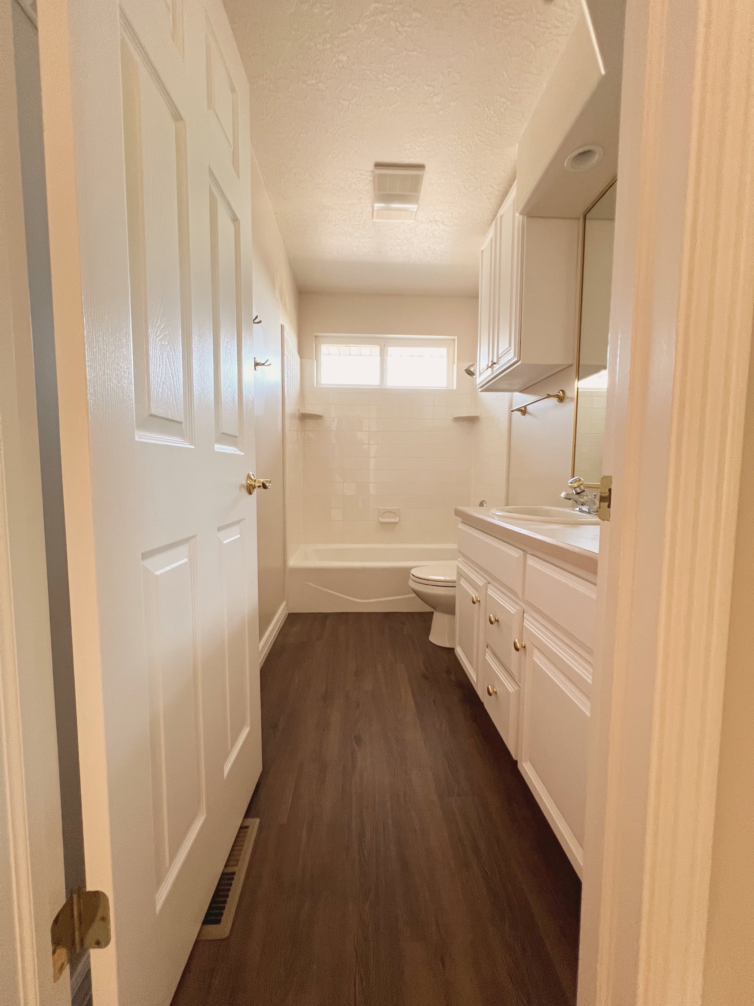 Bathroom featuring wood-finish flooring, a white vanity with gold-tone hardware, a built-in tub with overhead window, and white cabinetry