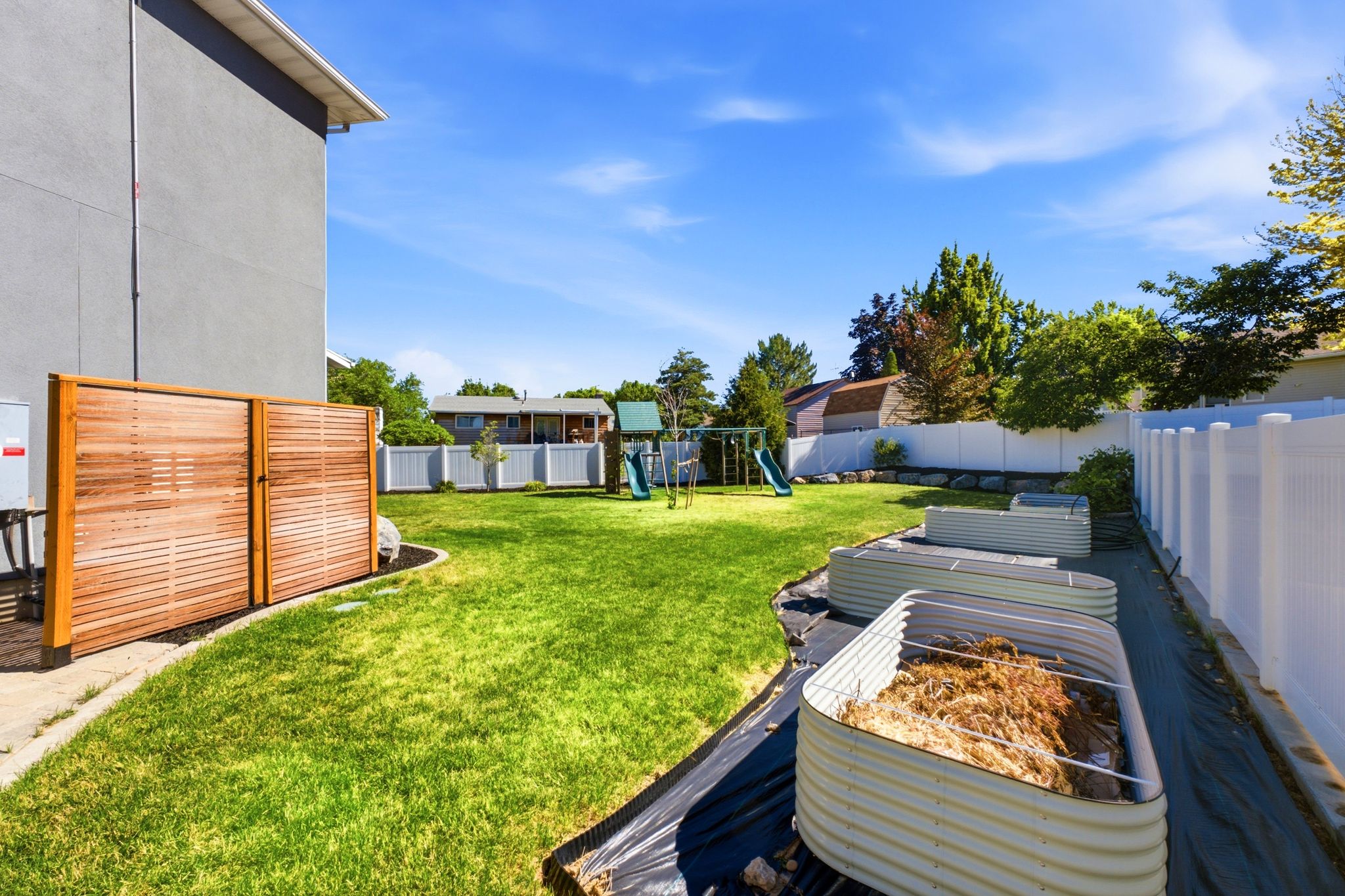 Expansive backyard featuring a manicured lawn, multiple raised garden beds, a white privacy fence, mature trees, and a wooden slatted screen