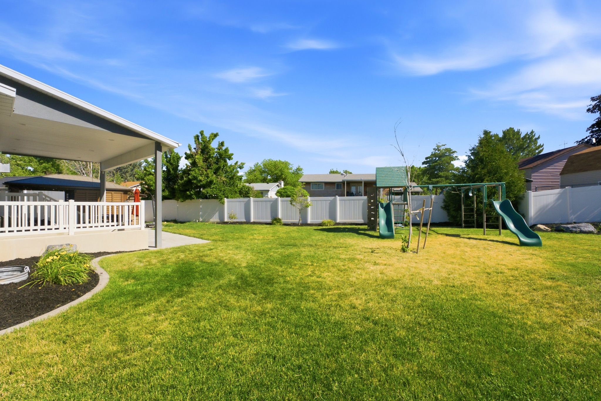 Expansive backyard featuring a covered patio with white railing, a concrete patio extension, a play structure with two slides, and a white vinyl privacy fence
