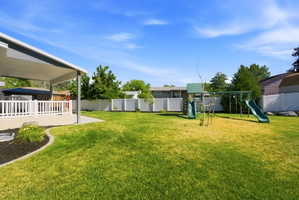 Expansive backyard featuring a covered patio with white railing, a concrete patio extension, a play structure with two slides, and a white vinyl privacy fence