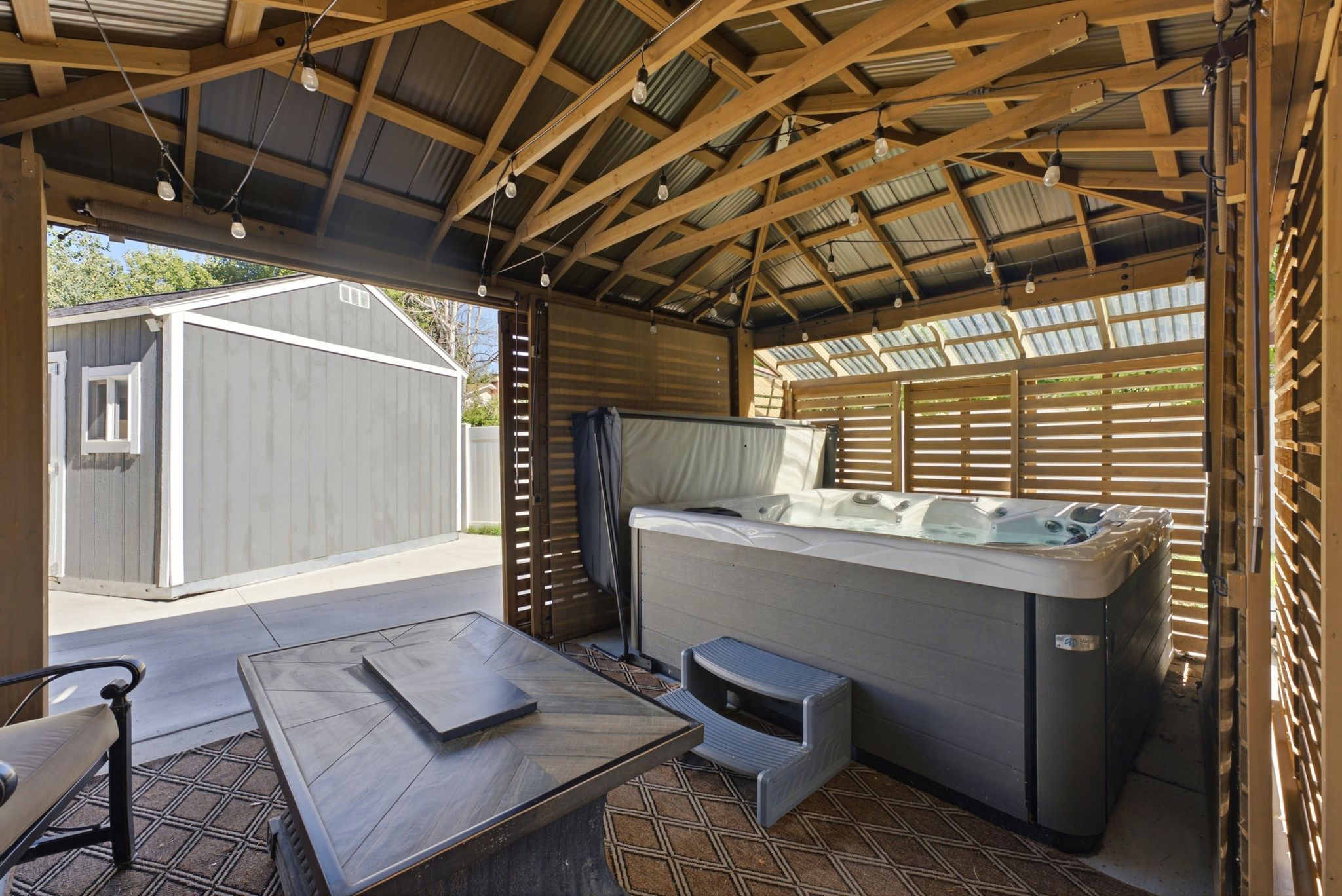 Covered outdoor spa with a wood-framed structure, corrugated metal roof, slatted wood privacy screens, and overhead string lighting