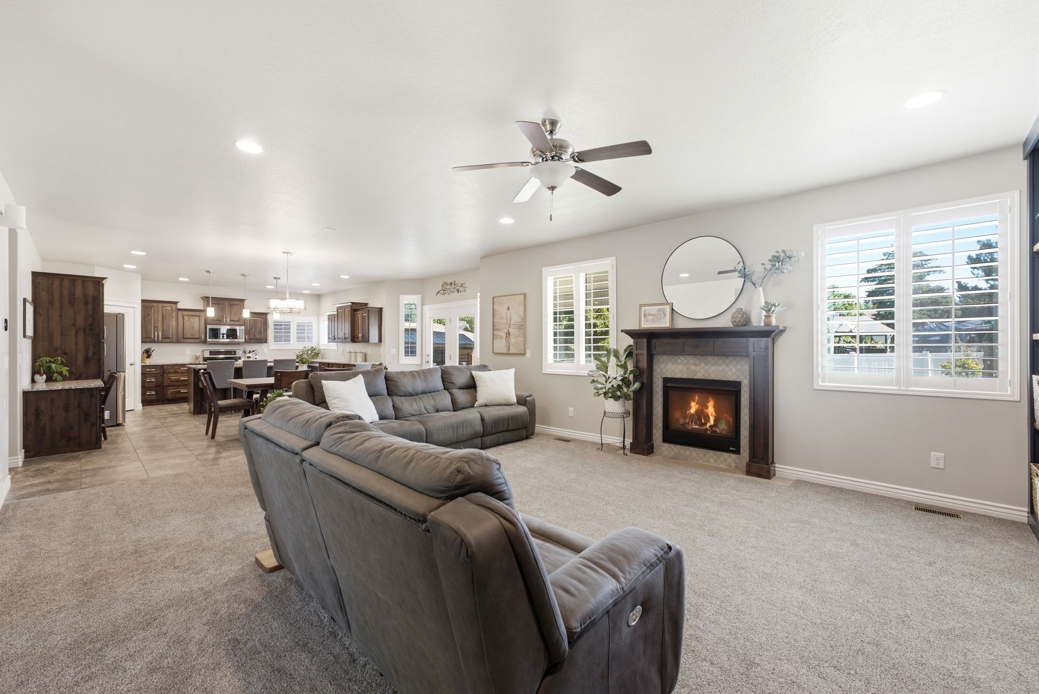 Open-concept living space featuring a fireplace with a dark wood mantel and tile surround, recessed lighting, ceiling fan, and multiple windows with plantation shutters
