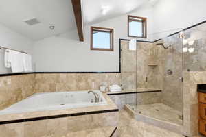 Spacious bathroom featuring a built-in jetted tub, a glass-enclosed shower with a built-in bench, natural stone tile flooring and wall surrounds, exposed wood ceiling beams, and dual high-set windows