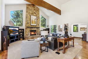 Spacious living area featuring a stone-clad fireplace, vaulted ceilings with exposed wood beams, large windows, wood-finish flooring, and light-toned carpeting