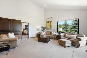 Spacious living area featuring a high vaulted ceiling, large picture window with tree scenery, light-toned carpeting, recessed lighting, and a built-in dark wood storage unit