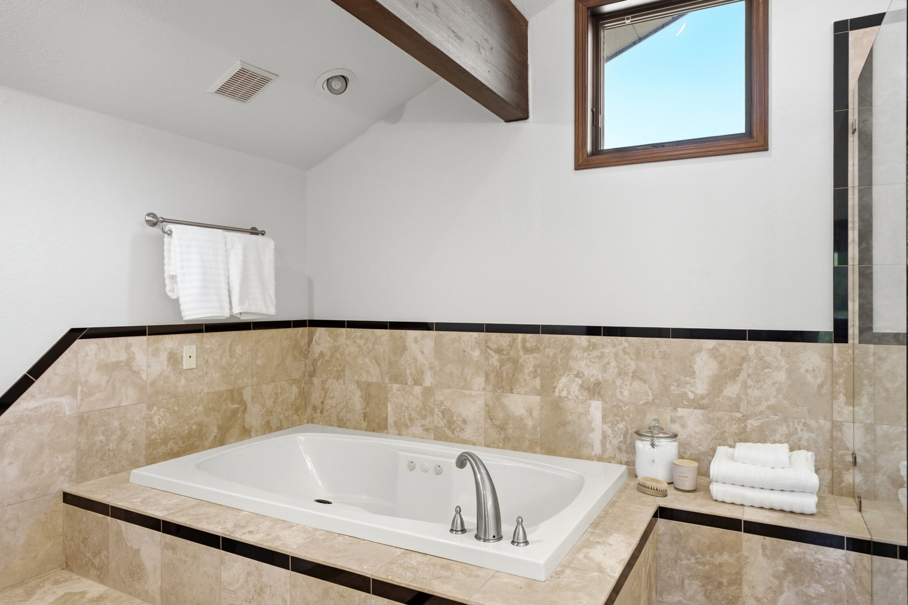 Built-in bathtub featuring a deck-mounted faucet and surround finished with light-toned stone tile and a dark accent stripe