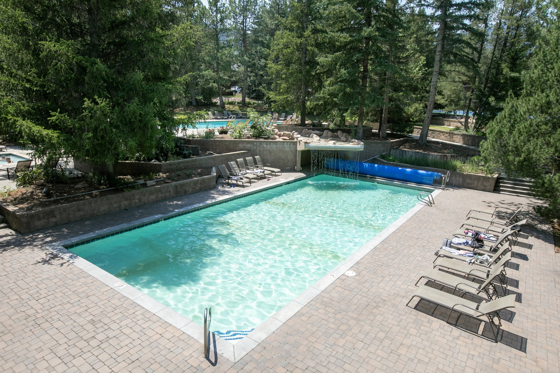 Outdoor swimming pool with brick paver surround, integrated waterfall feature, and tiered landscaping
