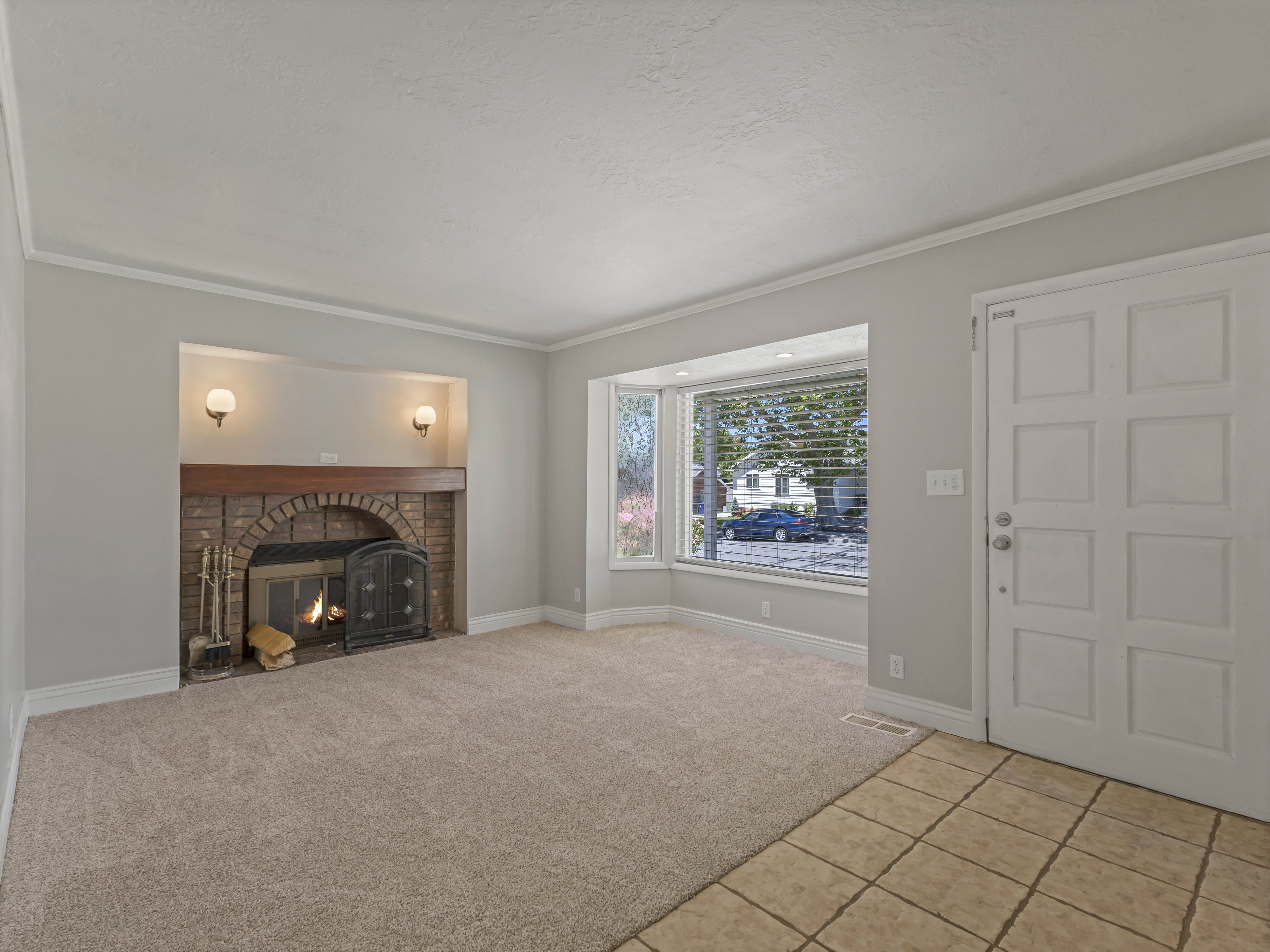 Spacious interior featuring a brick fireplace with sconce lighting, a multi-paneled entry door, a wide bay window with blinds, light-toned wall-to-wall carpeting, and tiled entry flooring