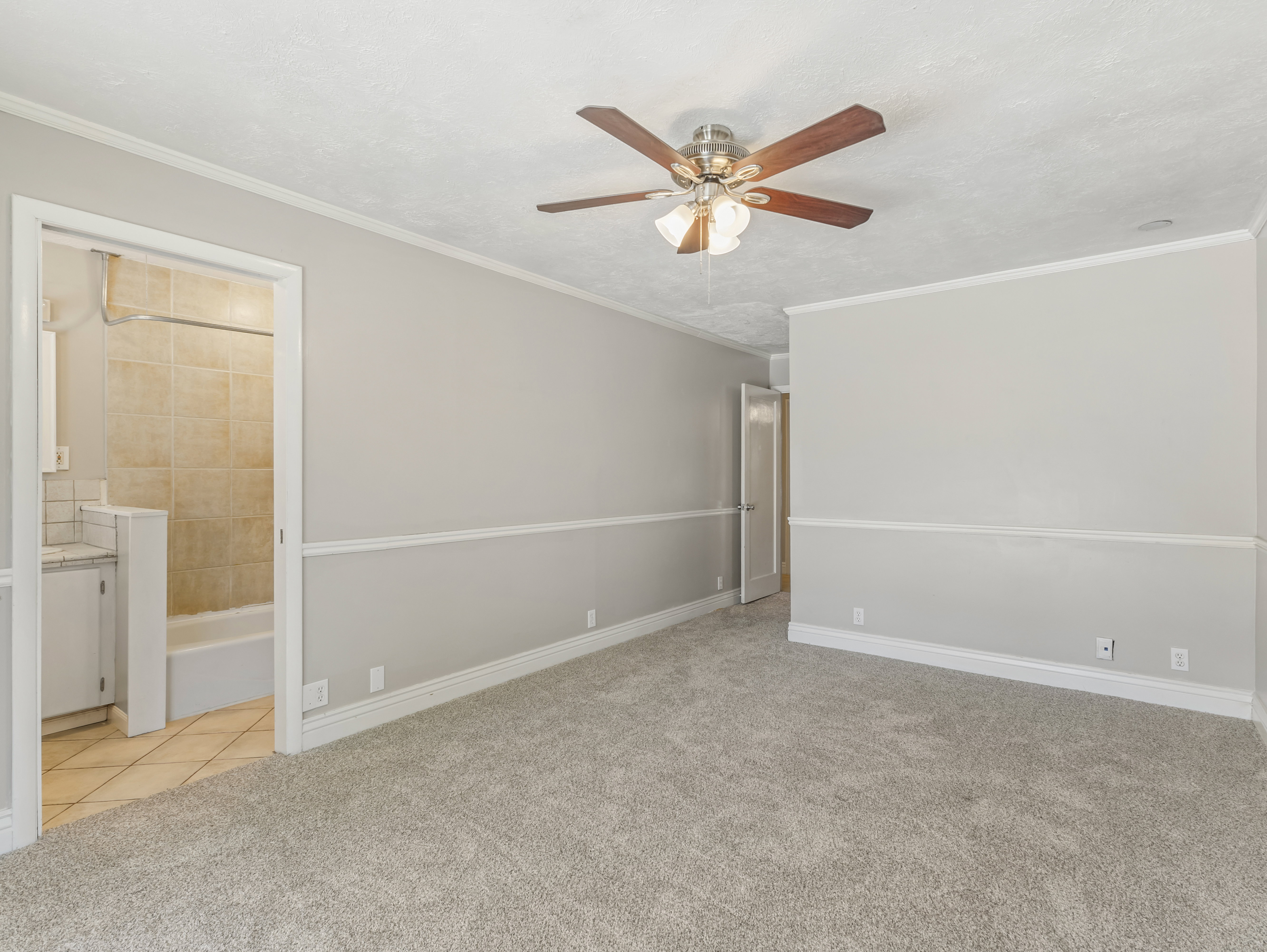 Spacious room featuring neutral carpeting, light gray walls with white chair rail molding, and a ceiling fan with integrated lighting