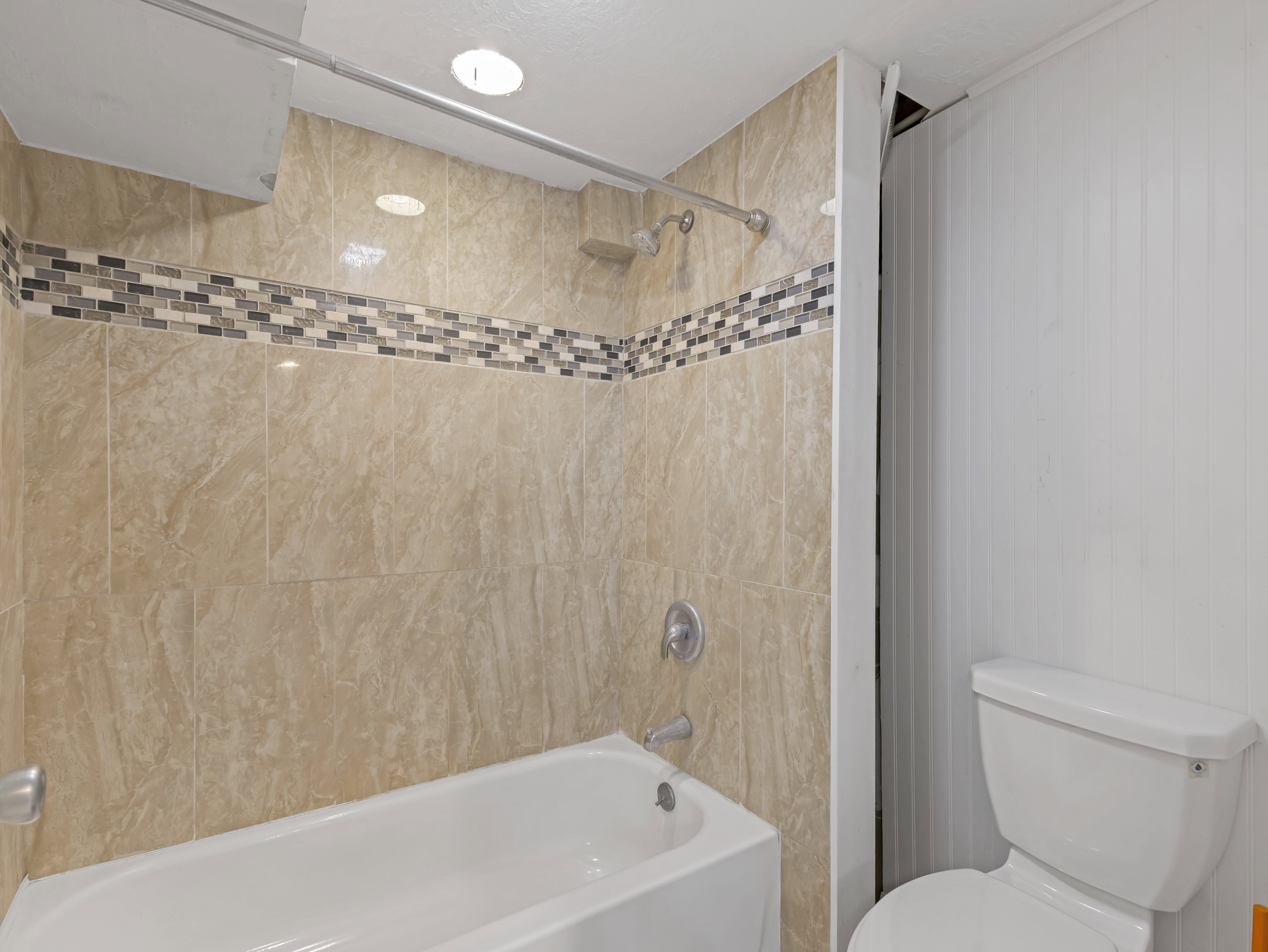 Bathroom featuring a white bathtub with a wall-mounted showerhead and faucet, tan ceramic wall tiling, a decorative mosaic tile accent band, and white beadboard paneling