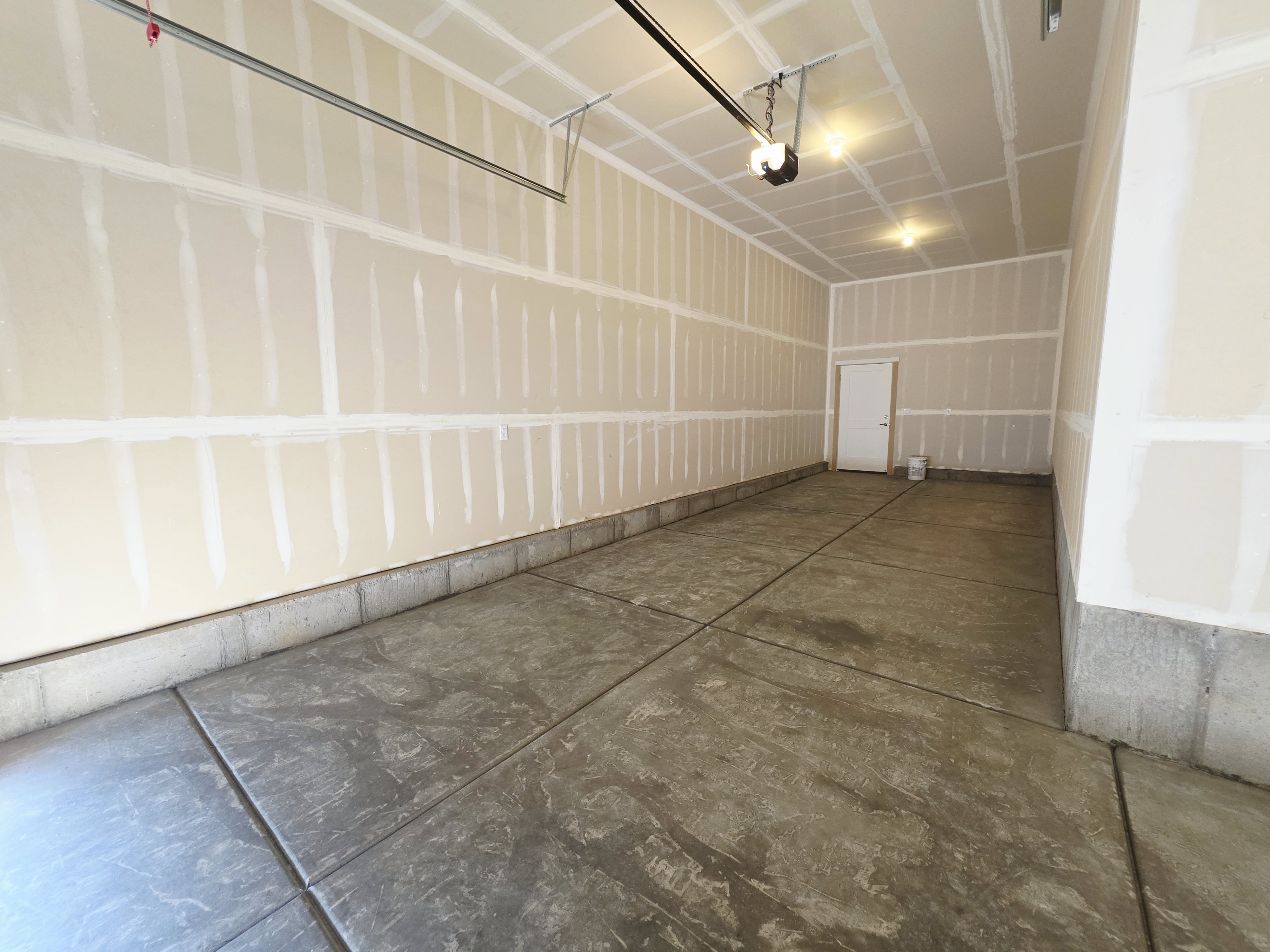 Spacious two-car garage featuring a concrete floor, exposed drywall walls, an overhead door mechanism, and ceiling-mounted lighting fixtures