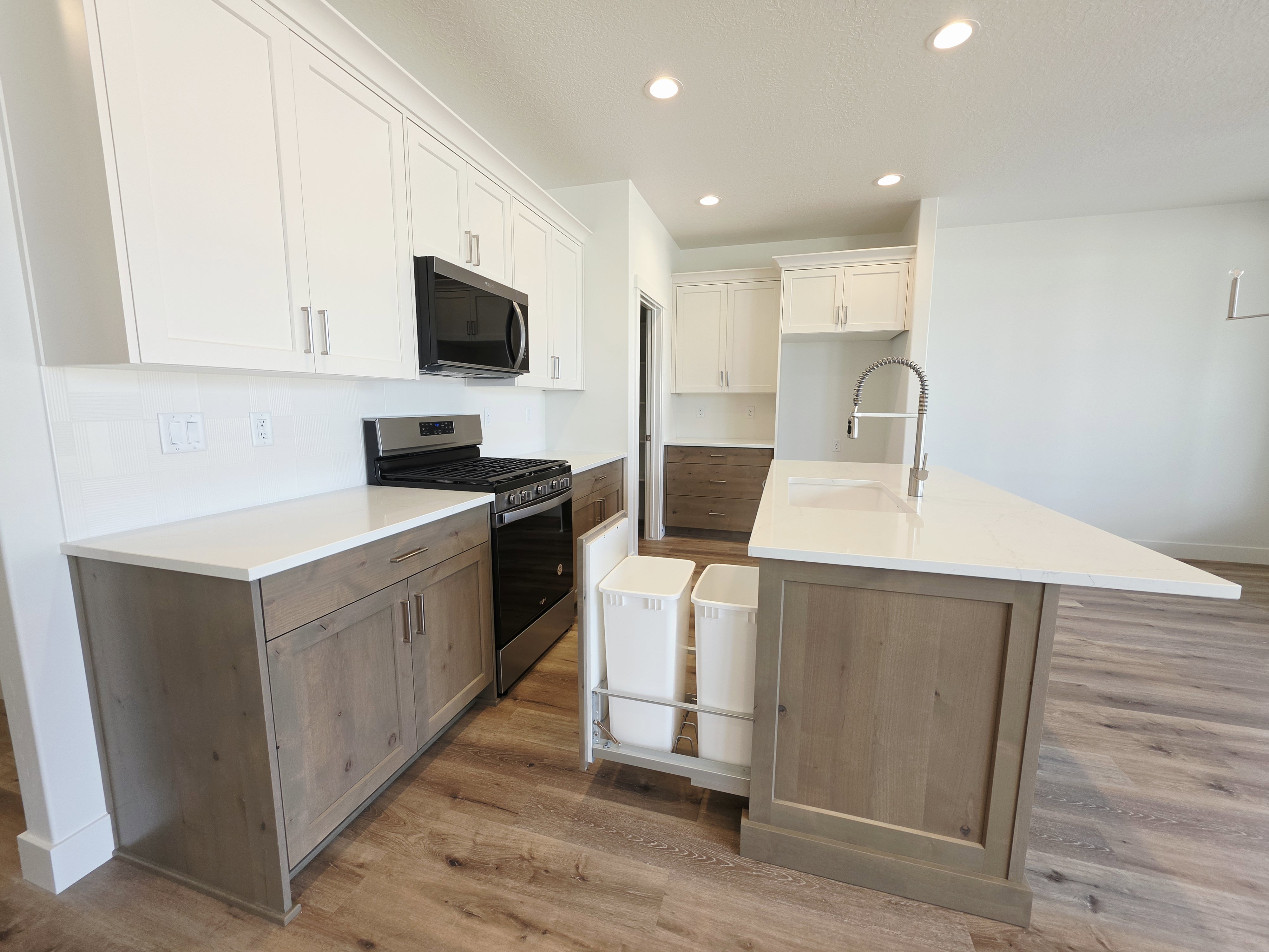 Kitchen featuring wood-finish flooring, two-tone cabinetry with white upper and wood-finish lower cabinets, white countertops, and a kitchen island with an integrated sink and pull-down faucet