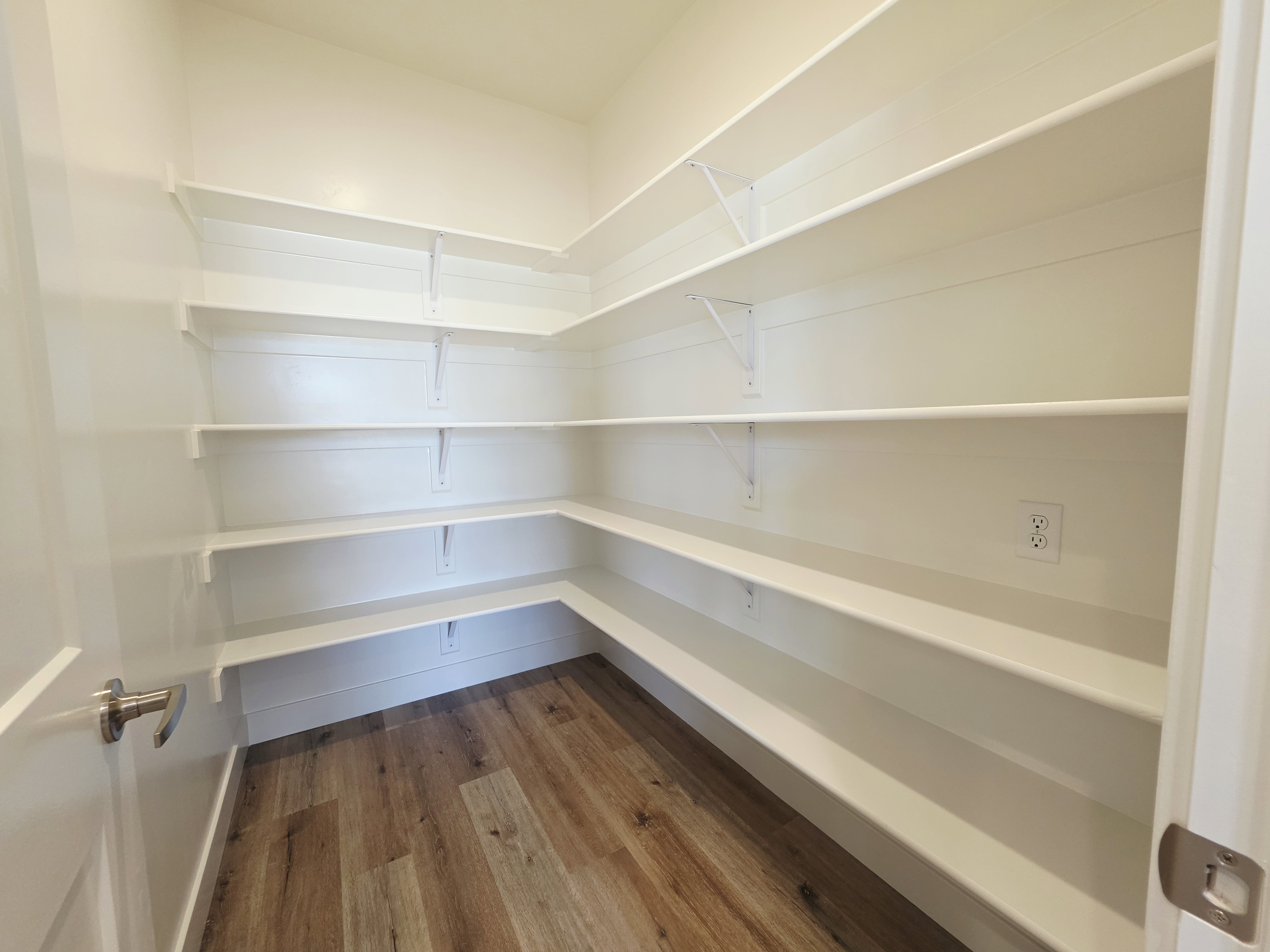 Walk-in pantry featuring extensive white shelving, wood-finish flooring, and an electrical outlet