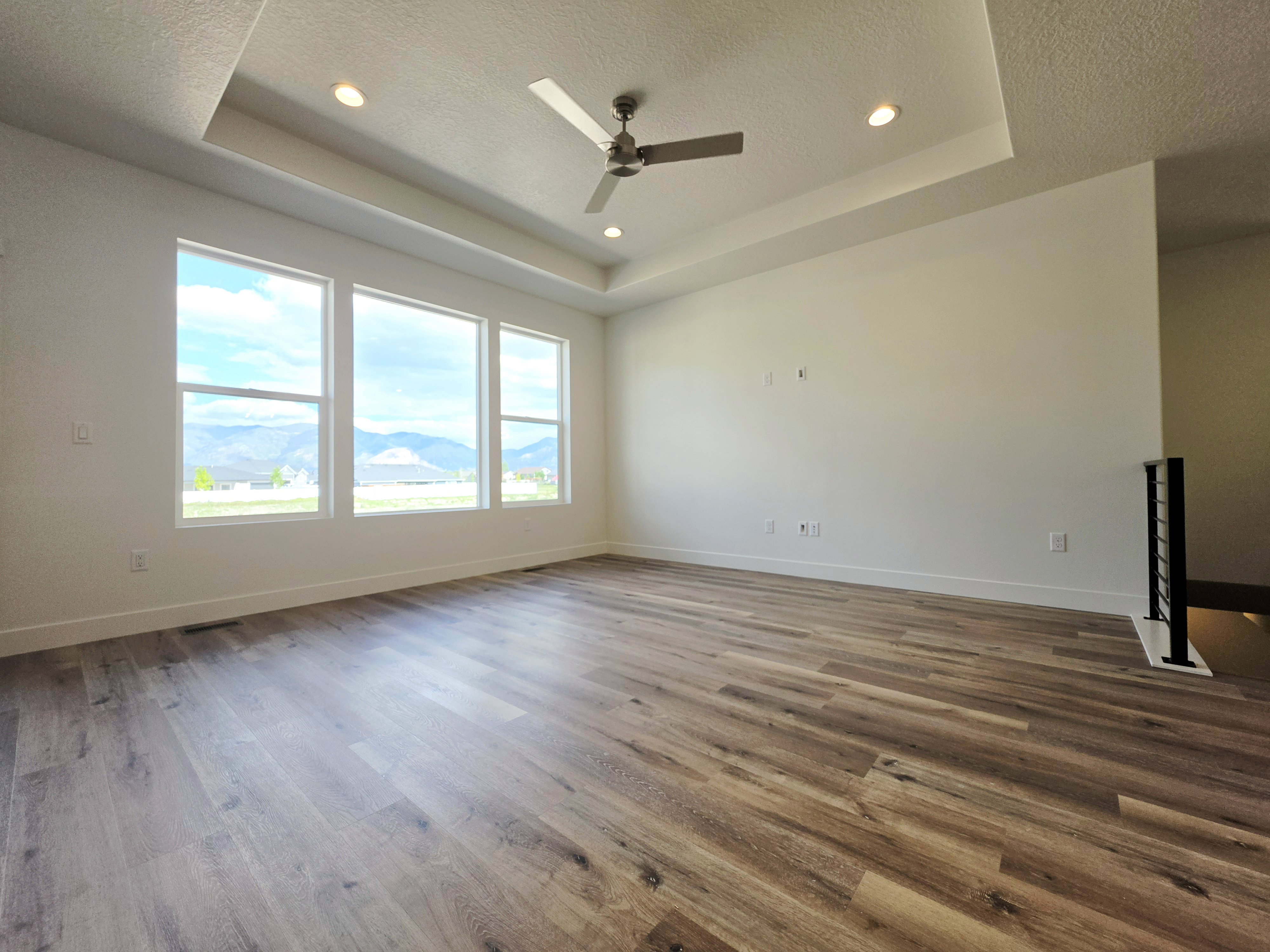 Spacious interior room featuring wood-finish flooring, a tray ceiling with recessed lighting and a ceiling fan, a wall of three large windows, and a black metal railing