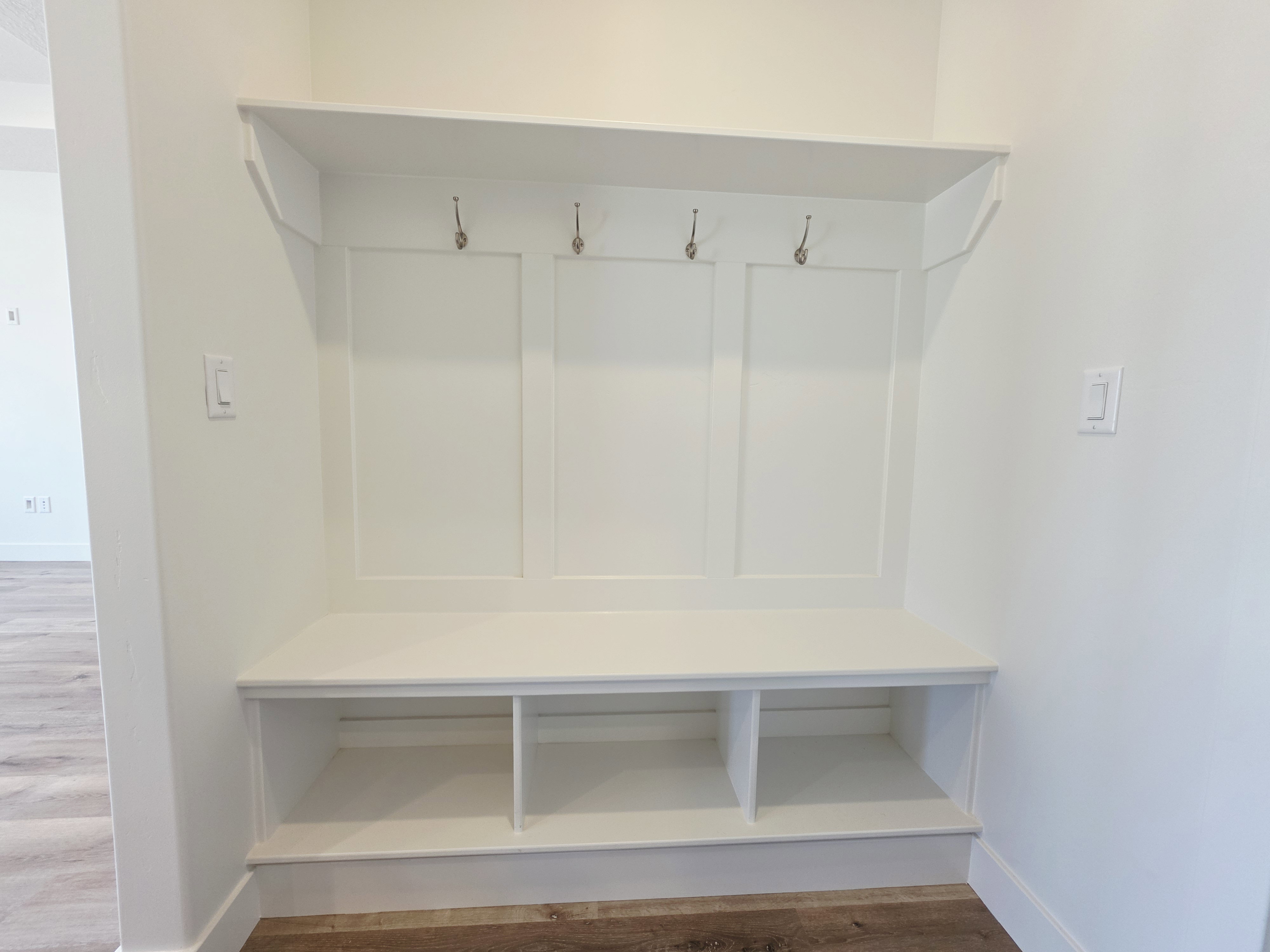 Built-in storage bench with three open compartments, three wall hooks, white paneling, and a top shelf
