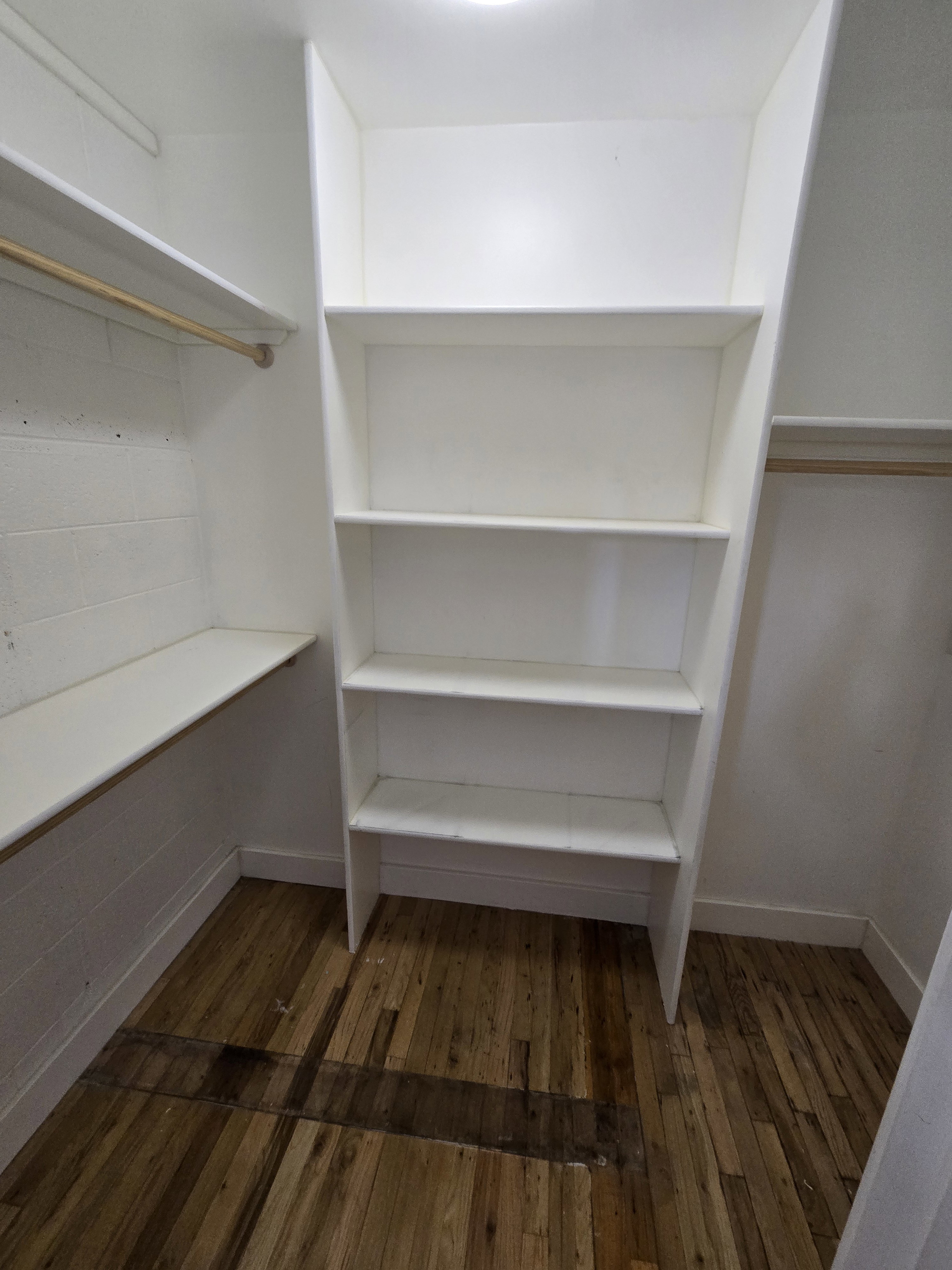 Built-in closet system featuring white shelving and a wooden hanging rod, complemented by wood-finish flooring
