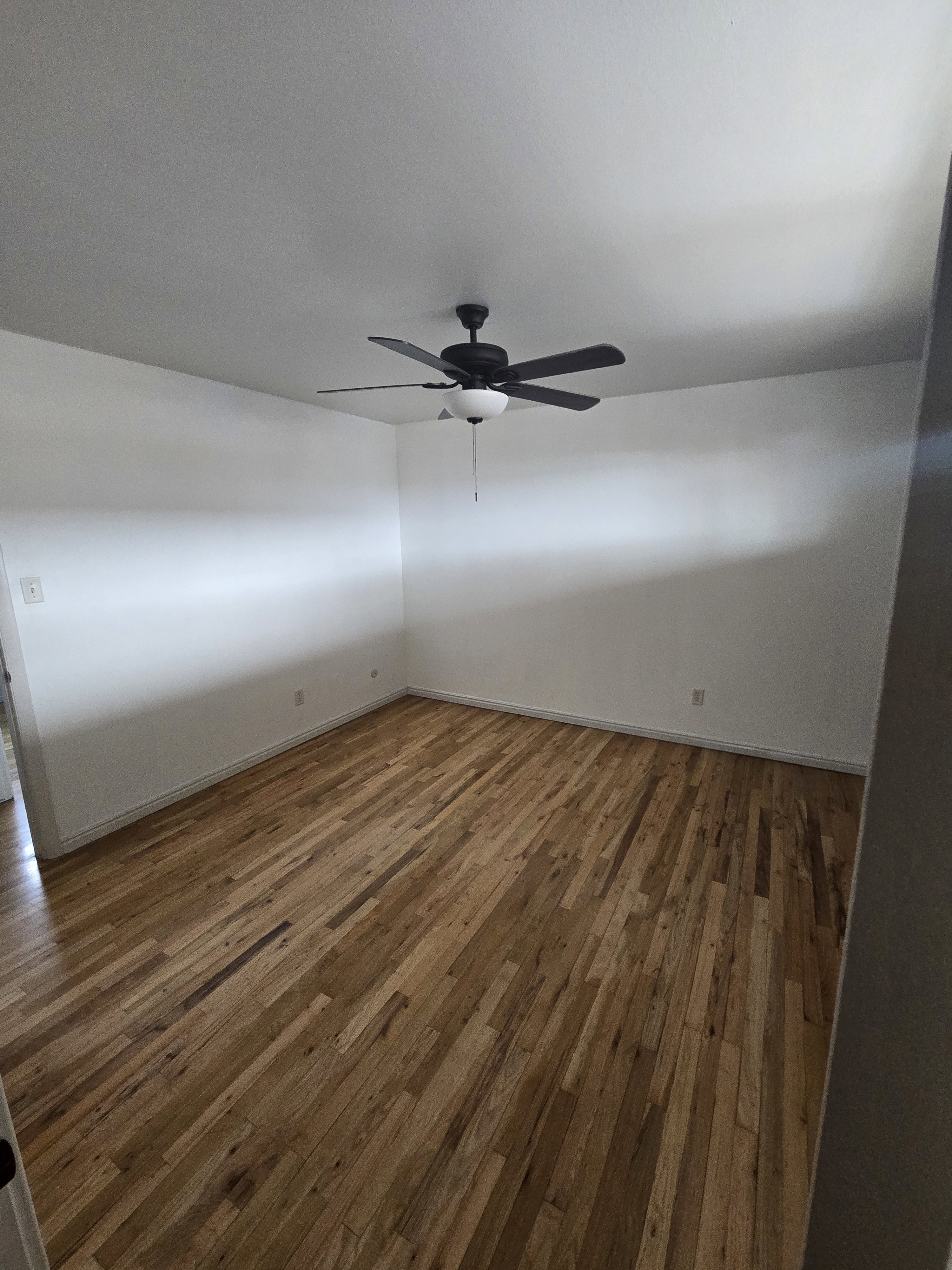 Spacious room with wood-finish flooring, white walls, and a ceiling fan with integrated lighting