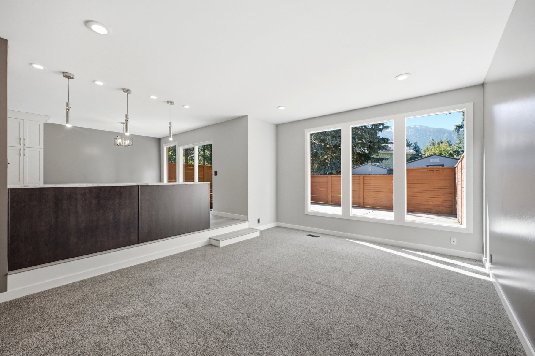Spacious living area featuring neutral carpeting, a partial wall with a dark wood-finish base and light countertop, recessed lighting, and three large windows overlooking a private fenced yard and distant mountains