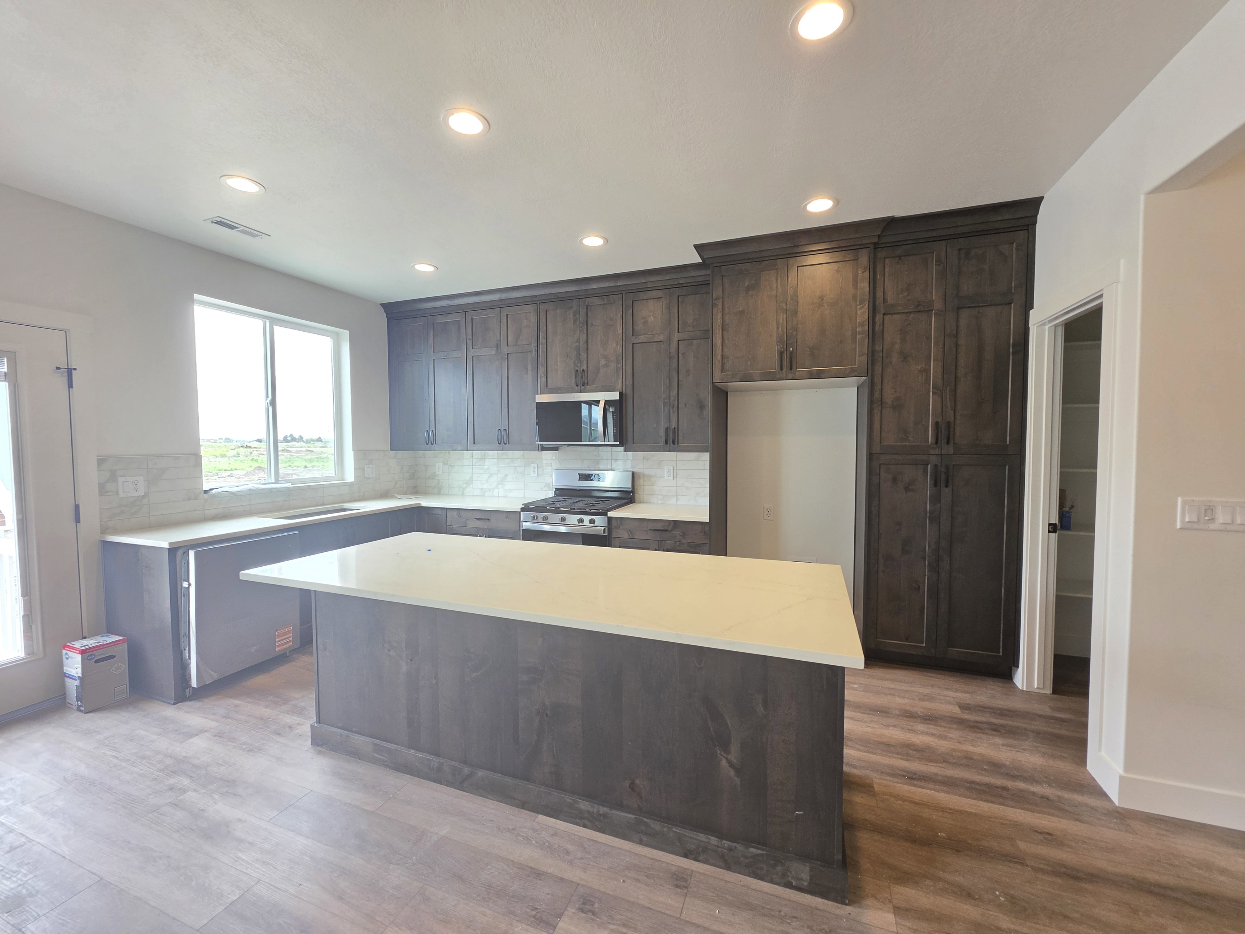 Modern kitchen featuring a large central island with a light-toned countertop, extensive dark wood cabinetry, stainless steel appliances, and wood-finish flooring