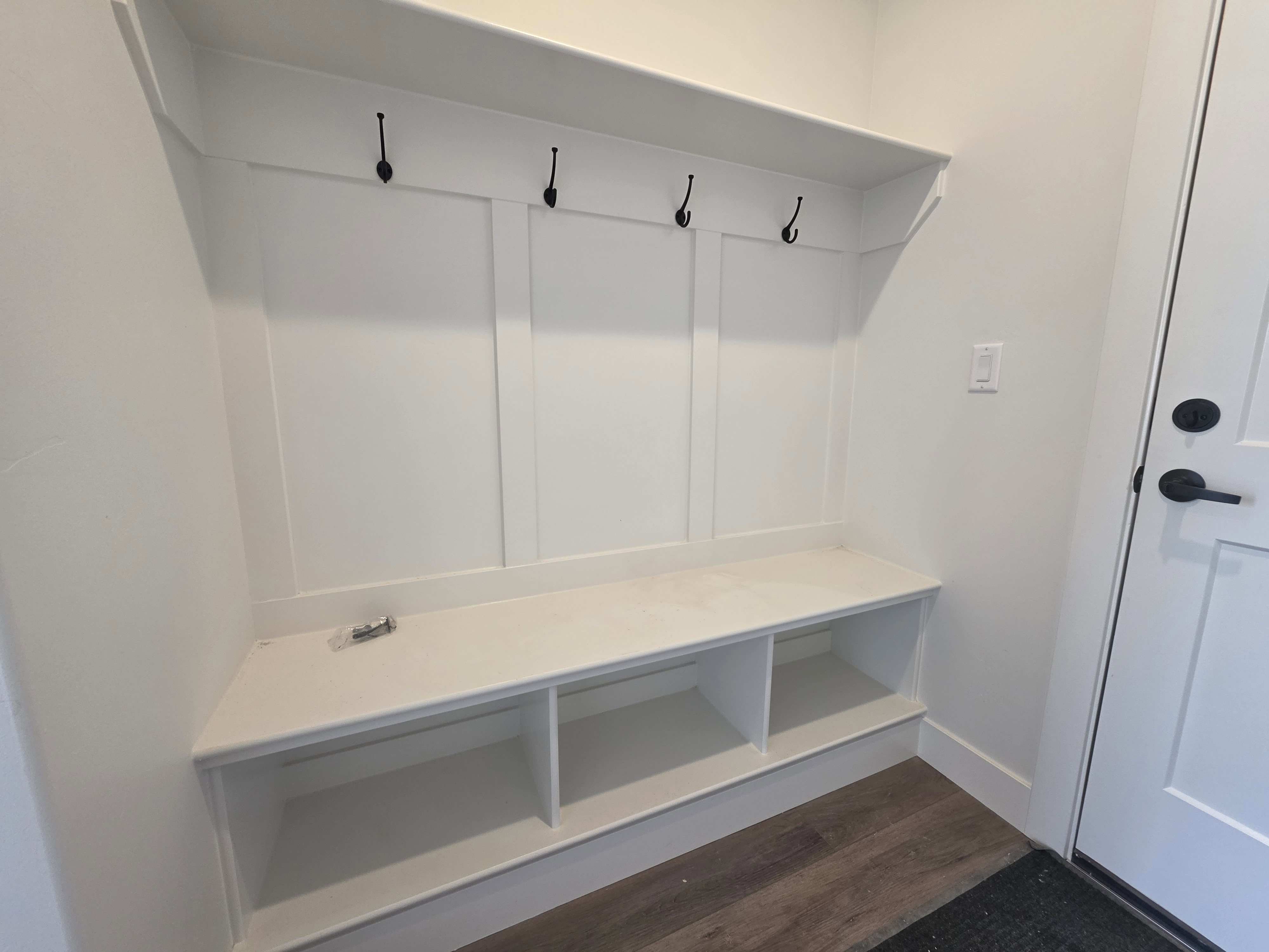 Built-in mudroom bench featuring three storage cubbies, a solid white countertop, and wainscoting with four black coat hooks