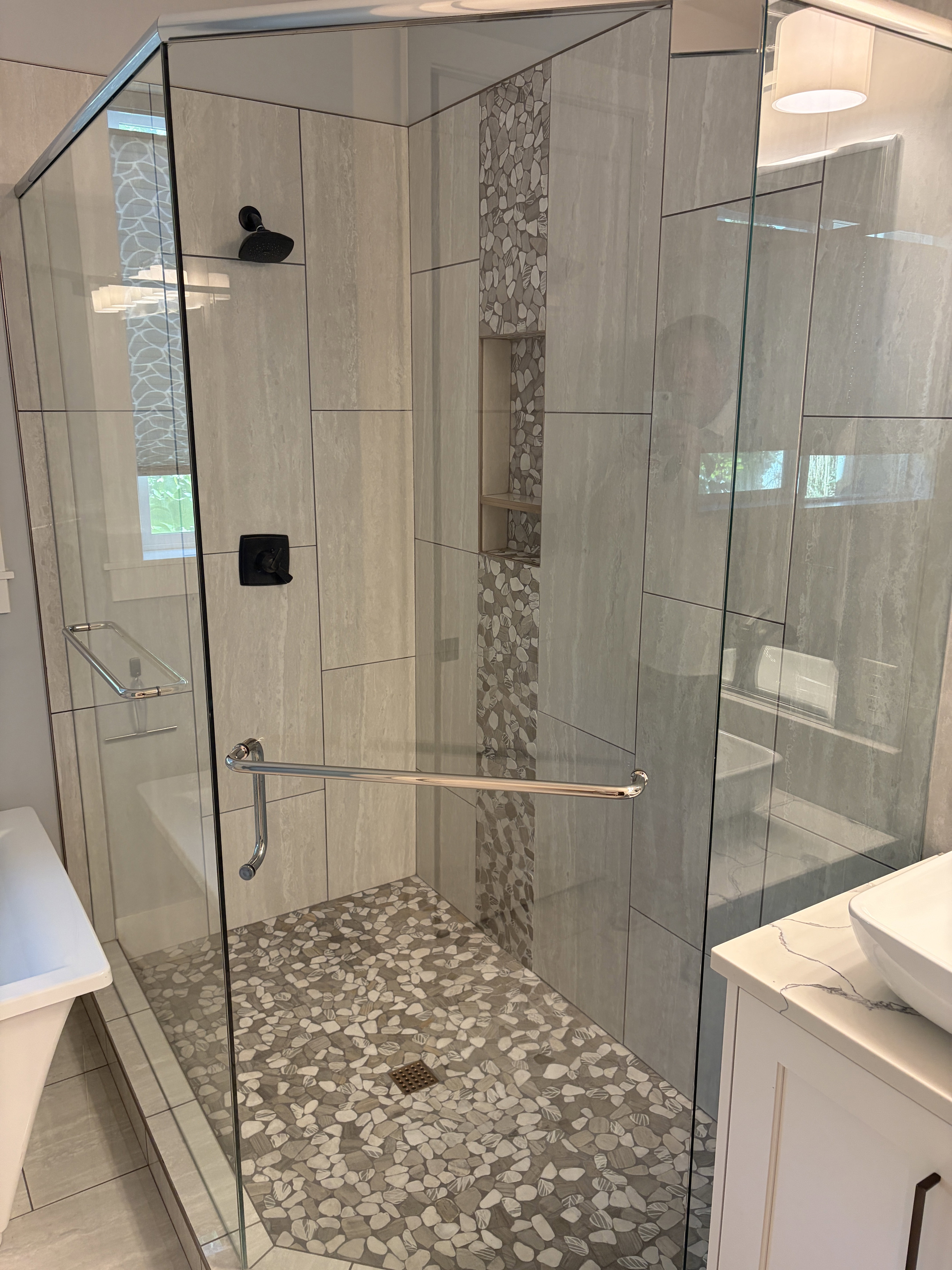 Glass-enclosed shower featuring a river rock floor, vertical tile accents, built-in shelving, a matte black shower head, and a coordinating matte black control
