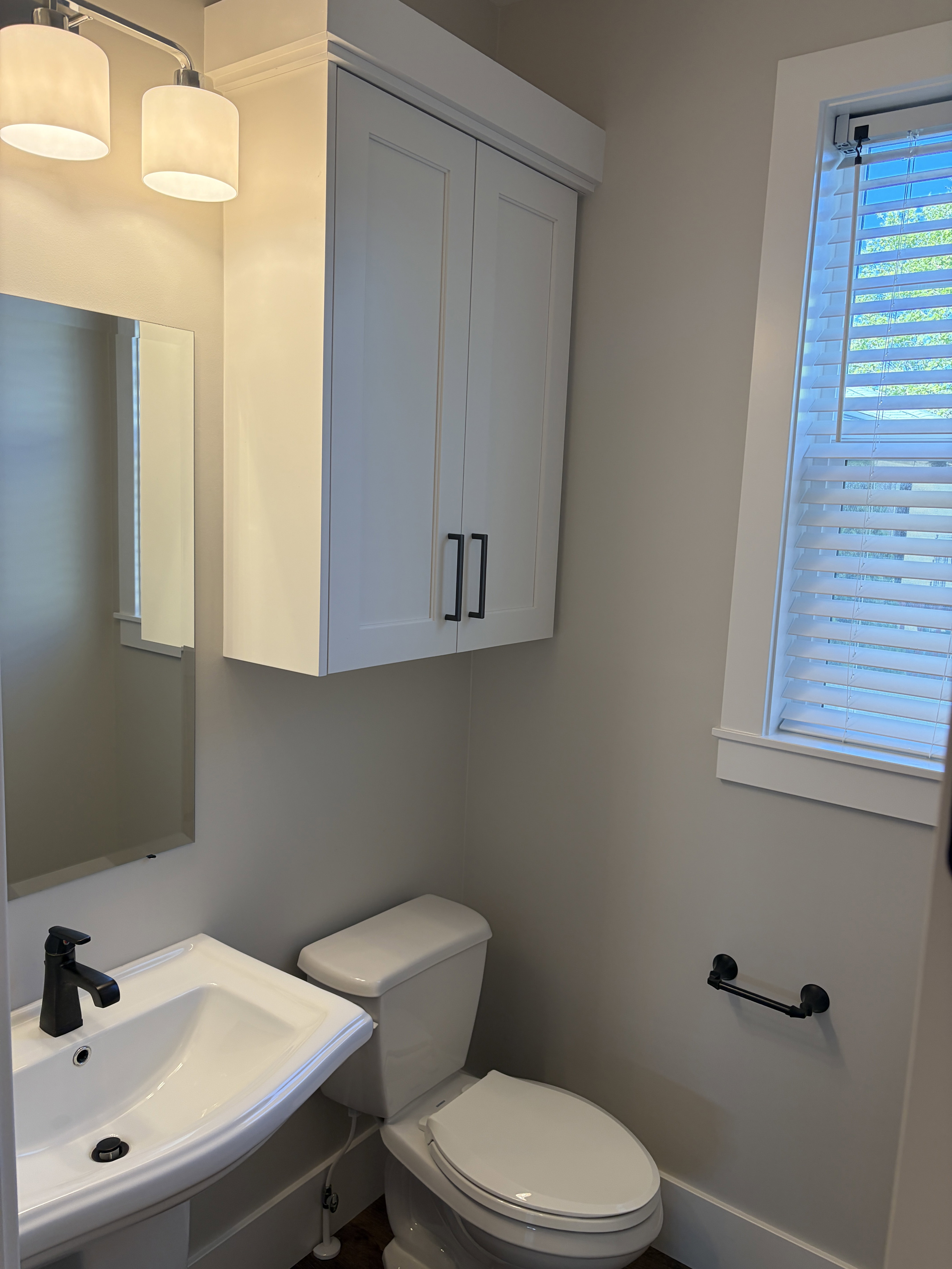 Pedestal sink with a matte black faucet, wall-mounted storage cabinetry, window with blinds, overhead vanity lighting, and wood-finish flooring