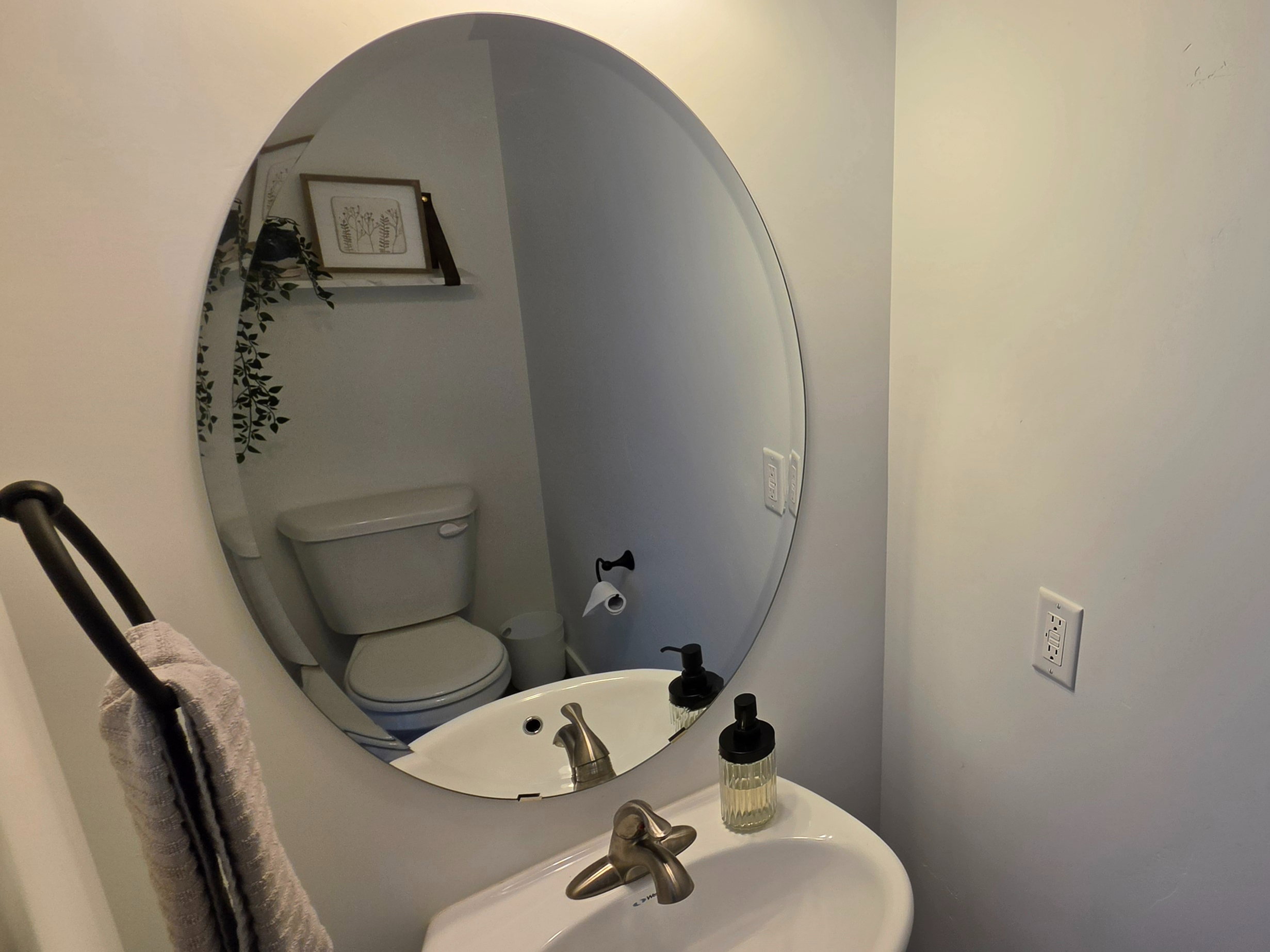 Pedestal sink with a brushed nickel faucet, large oval mirror, and a wall-mounted toilet