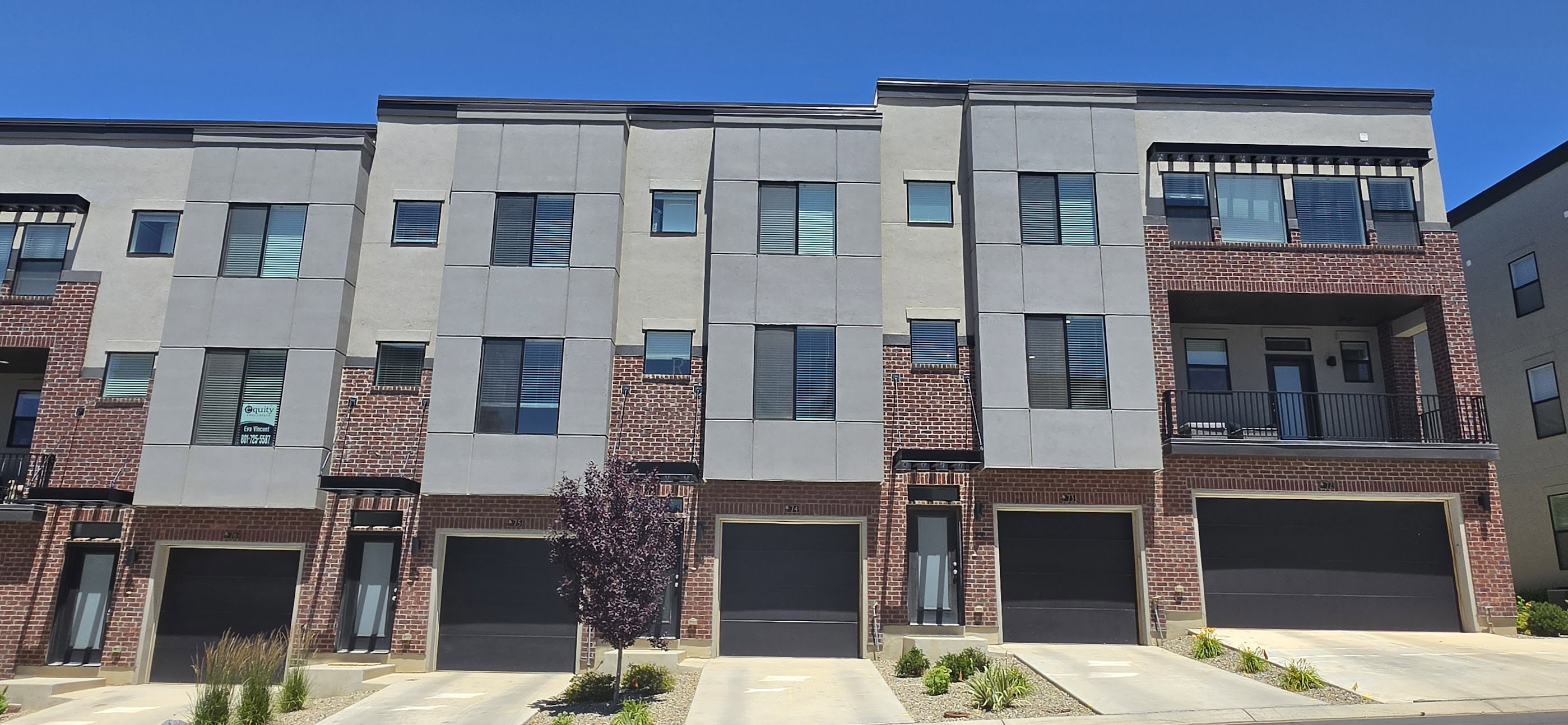 Modern townhome exteriors featuring red brick accents, light gray siding, black framed windows, and individual garage doors