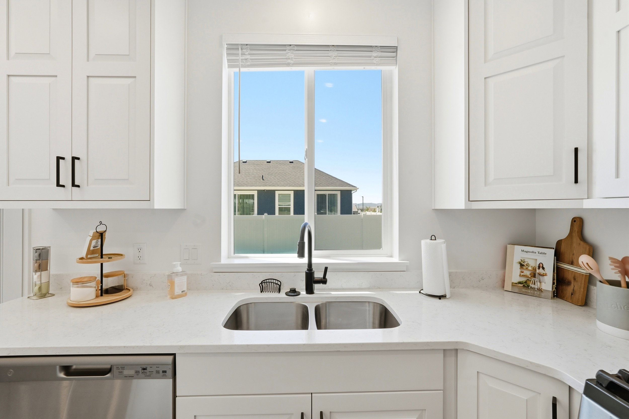 Kitchen featuring white shaker-style cabinetry with matte black hardware, light-toned countertops, a double basin stainless steel sink, and a black gooseneck faucet