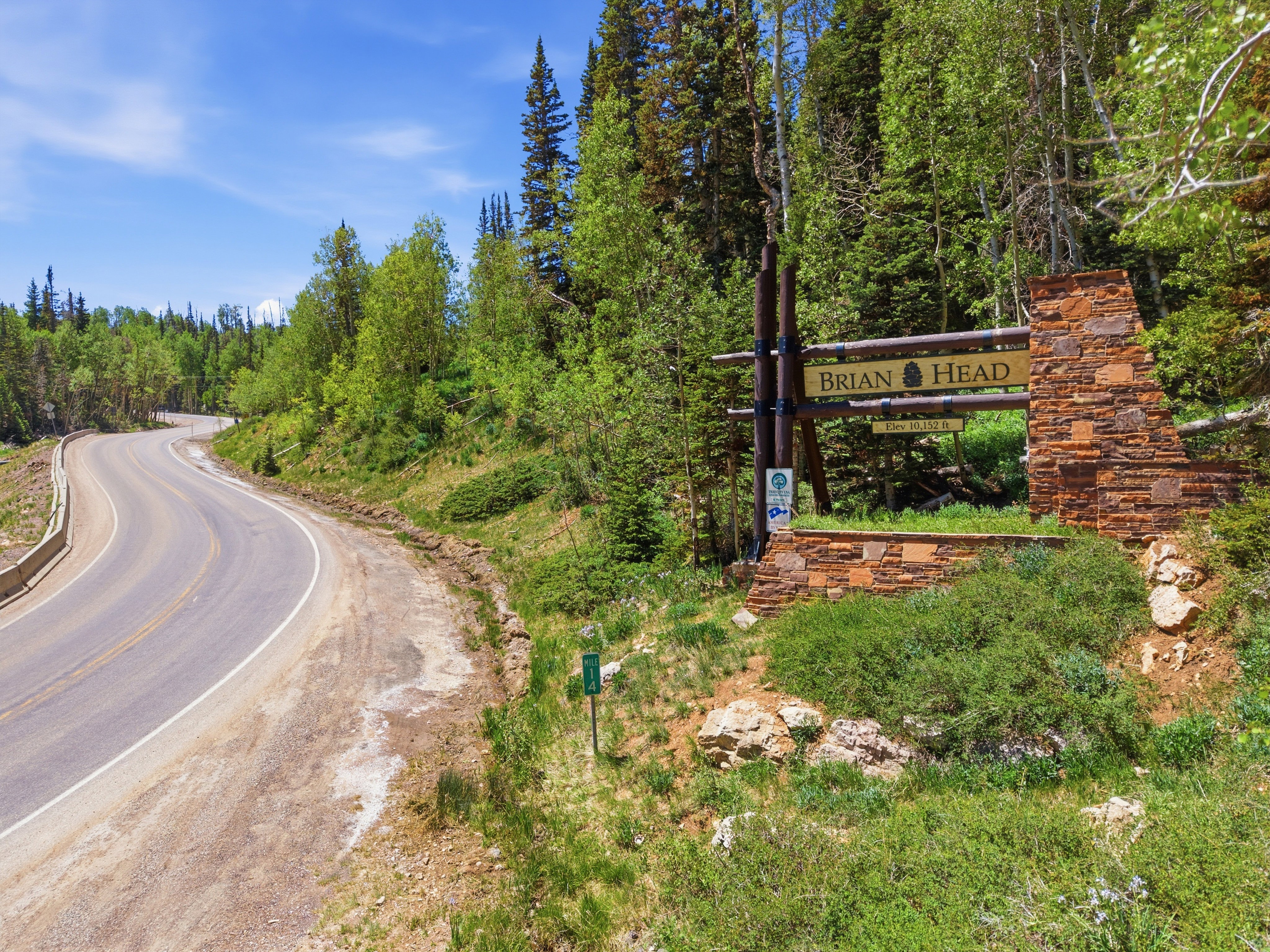Stone and timber entryway sign for Brian Head, set against a backdrop of mature trees and a curving paved road