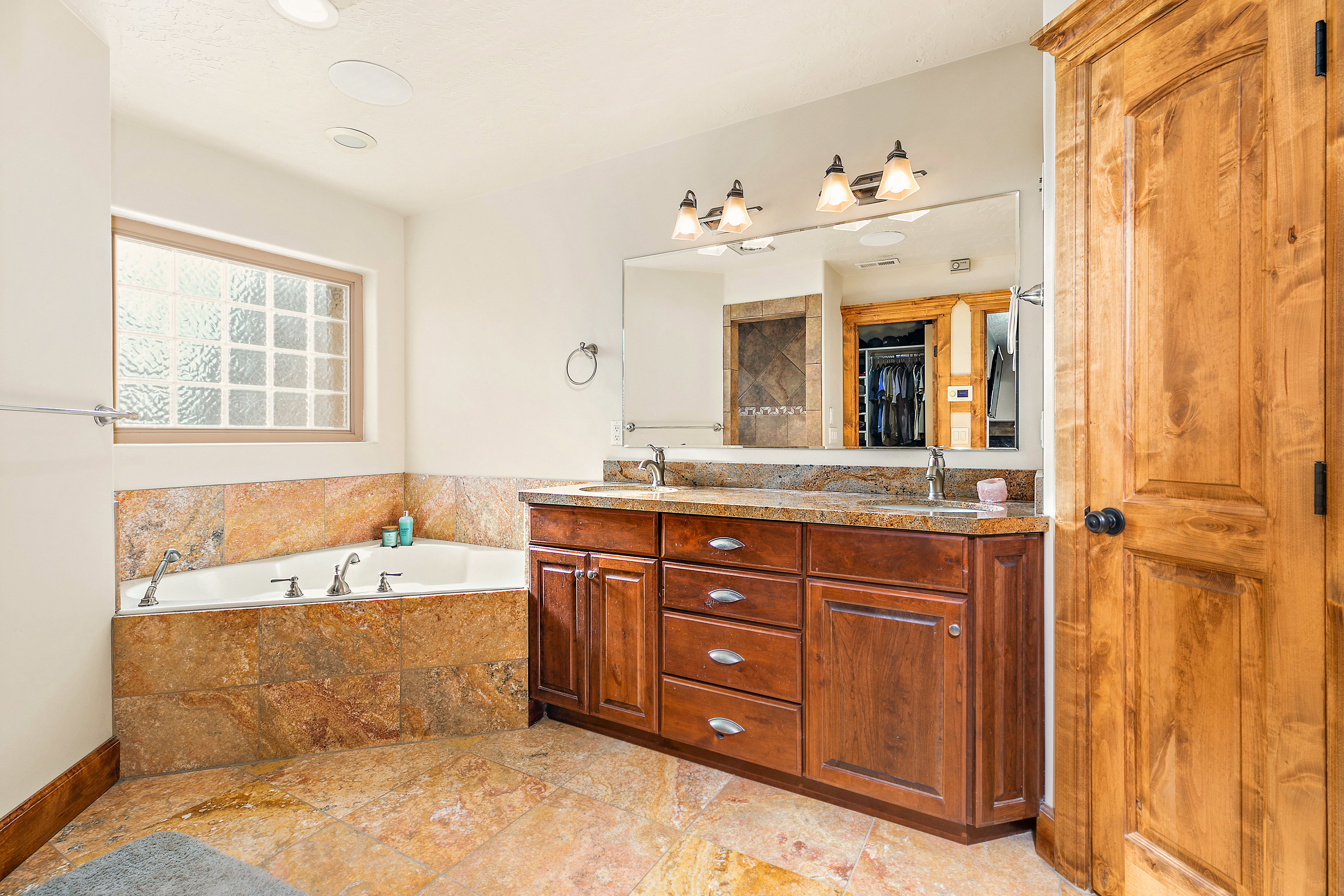 Spacious bathroom featuring a corner soaking tub with stone tile surround, a dual-sink wood vanity with granite countertop, and stone tile flooring
