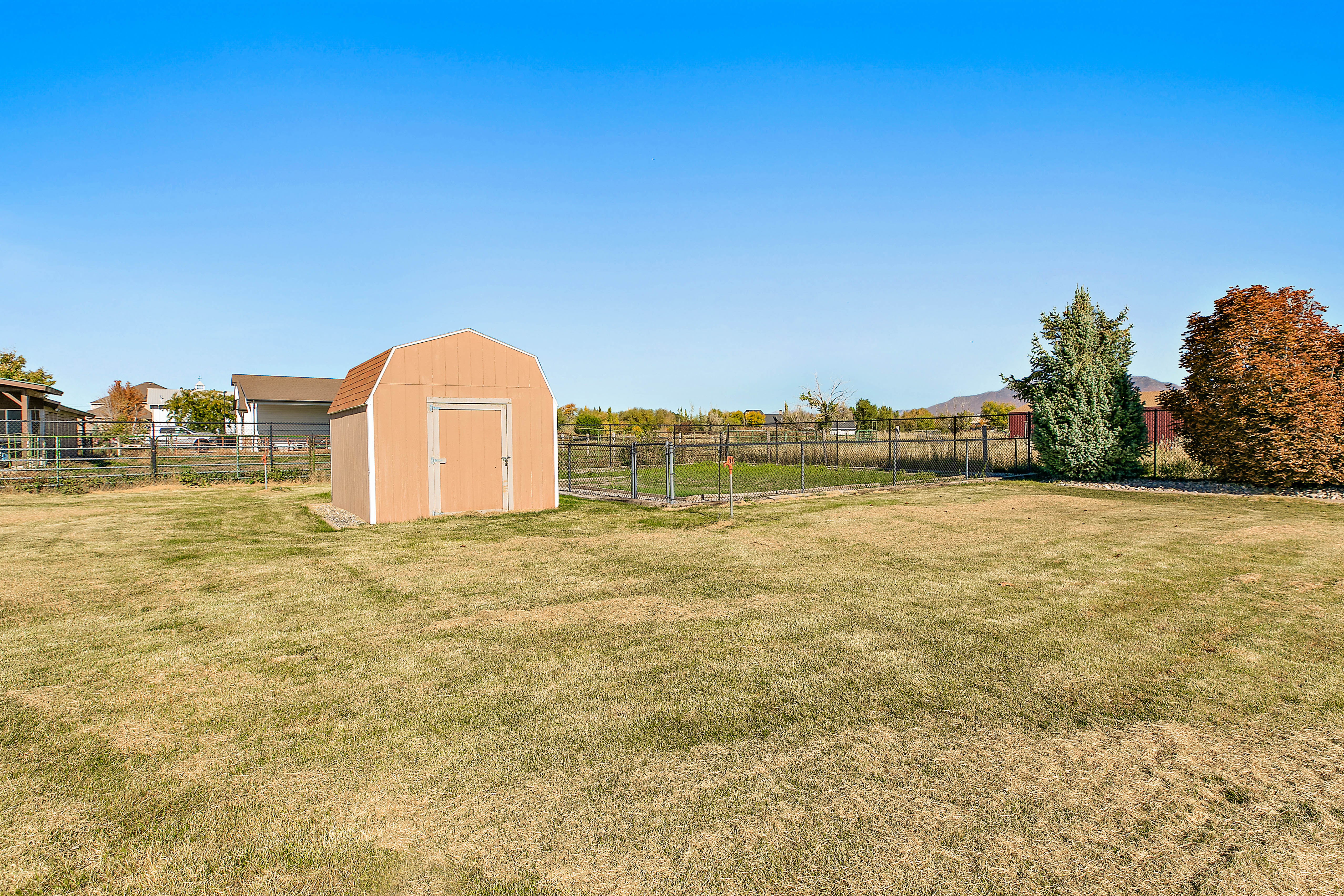 Expansive grassy yard featuring a light brown storage shed with a gambrel roof, chain-link fencing, and mature trees