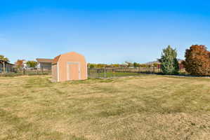 Expansive grassy yard featuring a light brown storage shed with a gambrel roof, chain-link fencing, and mature trees