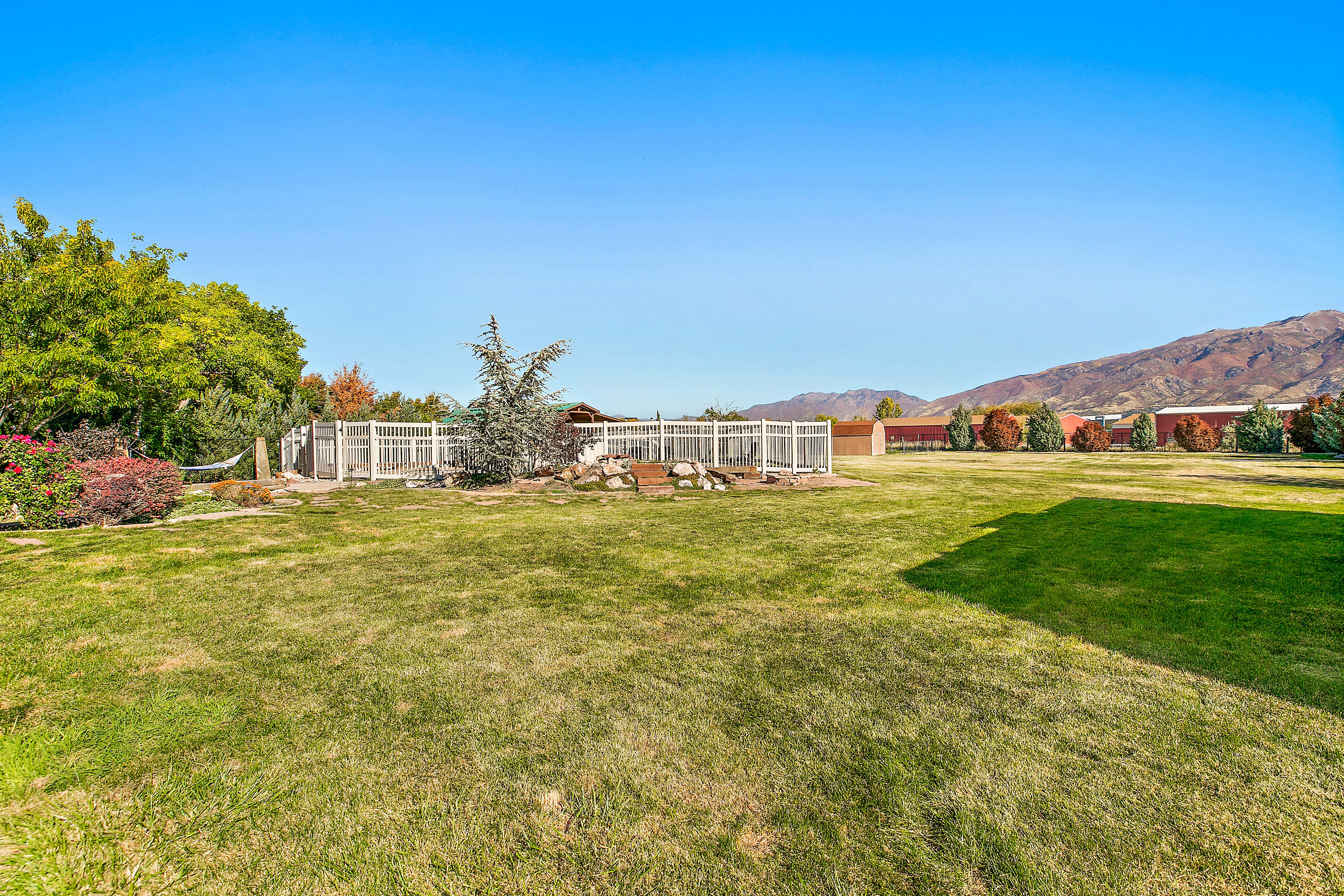Expansive grassy yard with mountain backdrop, surrounded by mature trees and shrubs, featuring white fencing and a dedicated enclosed area