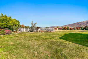 Expansive grassy yard with mountain backdrop, surrounded by mature trees and shrubs, featuring white fencing and a dedicated enclosed area