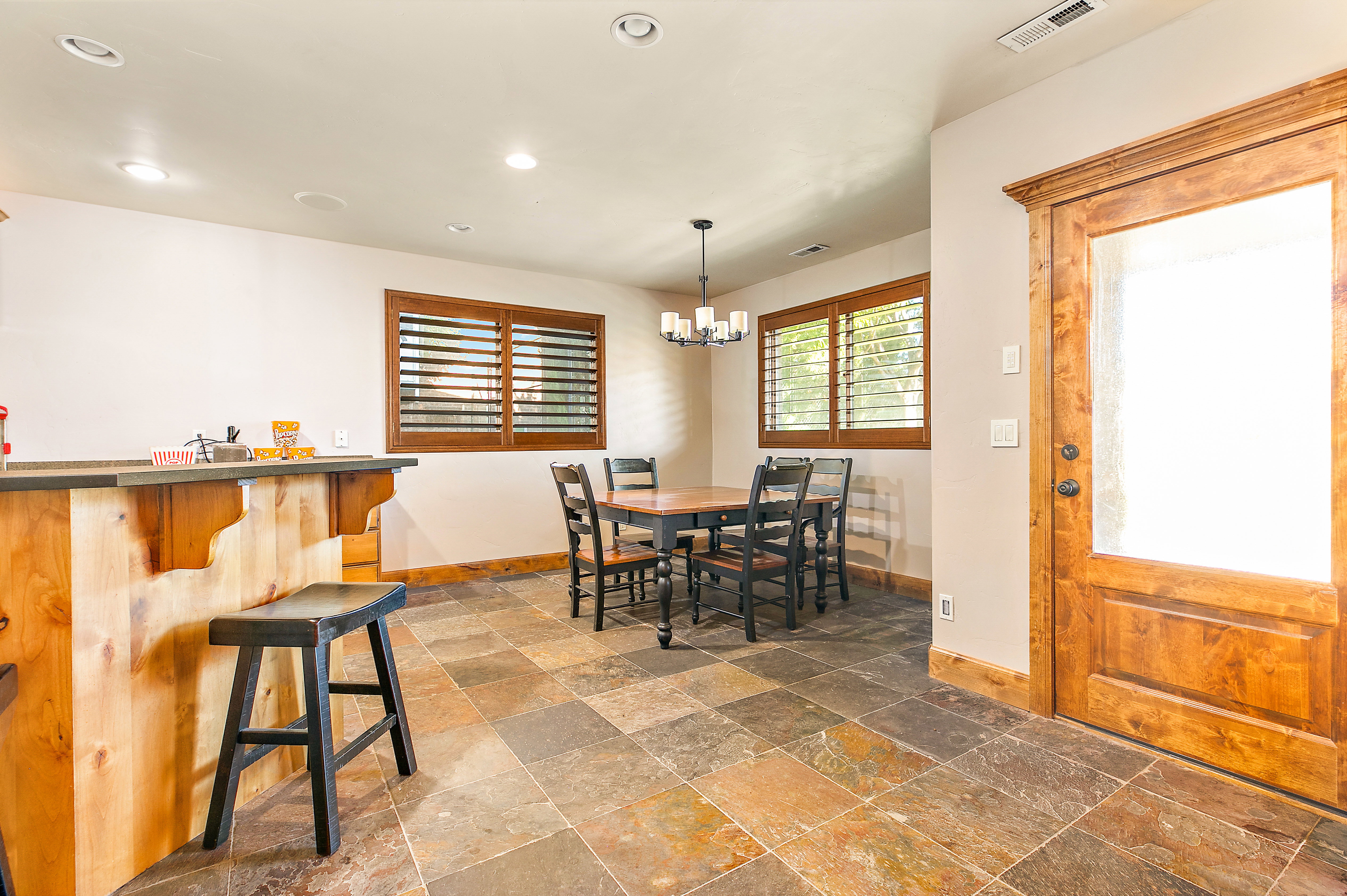 Spacious recreation area featuring multi-toned slate tile flooring, a wood-paneled bar, wood plantation shutters, and a wood entry door with opaque glass