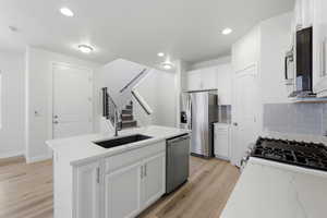 Modern kitchen featuring an island with an undermount sink and a stainless steel faucet, light wood-finish flooring, white cabinetry, stainless steel appliances, and a light blue subway tile backsplash