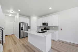 Modern kitchen featuring a central island with an undermount sink, stainless steel appliances, white cabinetry, and light wood-finish flooring