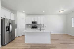 Open-concept kitchen featuring an island with a built-in sink, stainless steel appliances, white cabinetry, and light wood-finish flooring