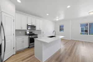 Kitchen featuring white shaker-style cabinetry, stainless steel appliances, a light grey subway tile backsplash, an island with a black undermount sink and gooseneck faucet, and wood-finish flooring