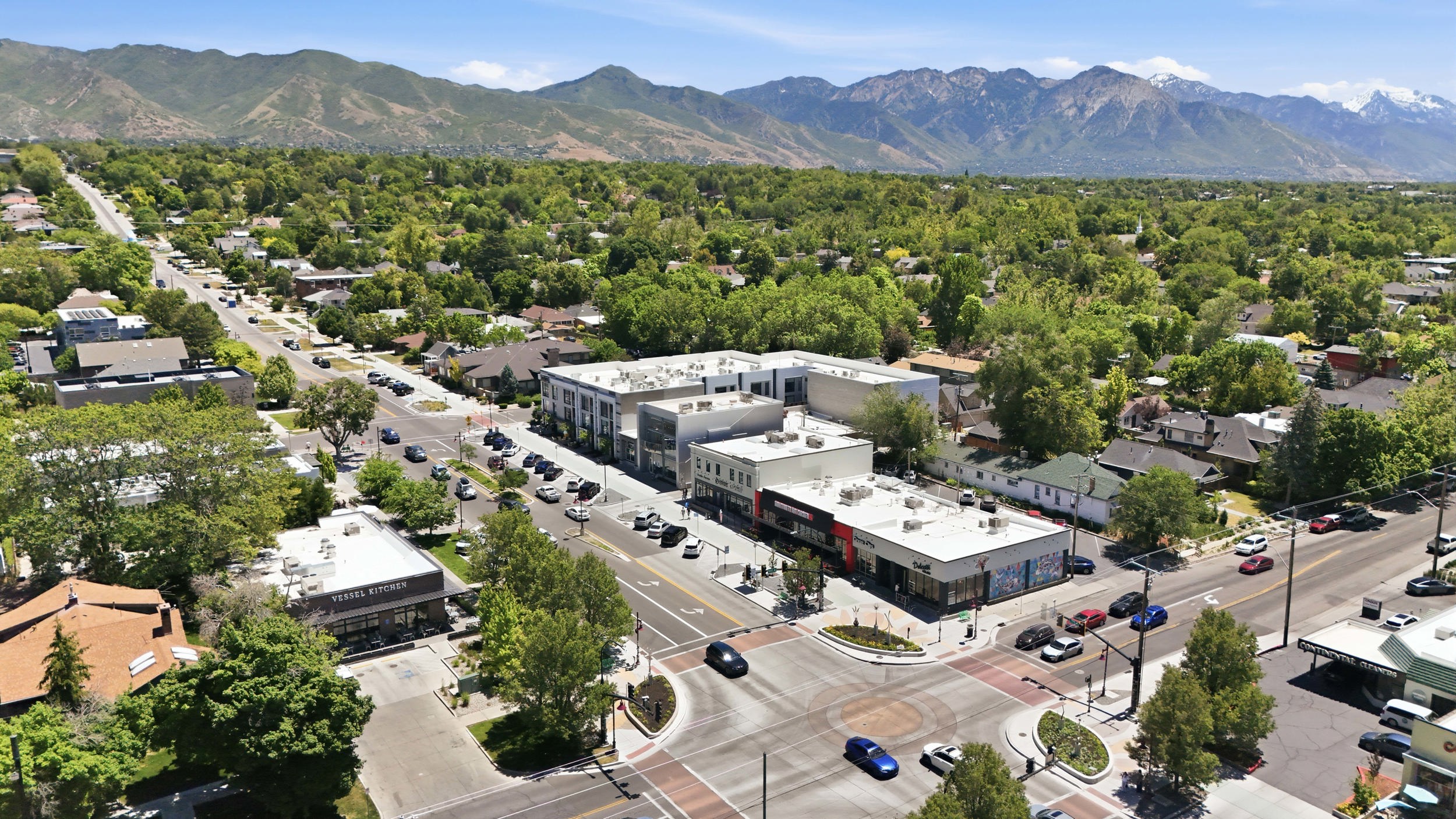 Aerial perspective of a commercial intersection with multiple businesses and residential properties, framed by distant mountains and abundant green trees