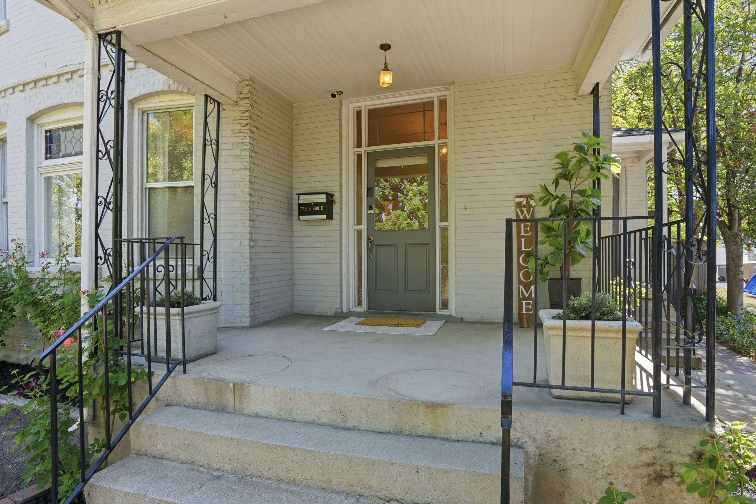 Inviting front porch featuring a sage green entry door with sidelights, a white brick facade, concrete steps, wrought iron railings, and a hanging pendant light fixture