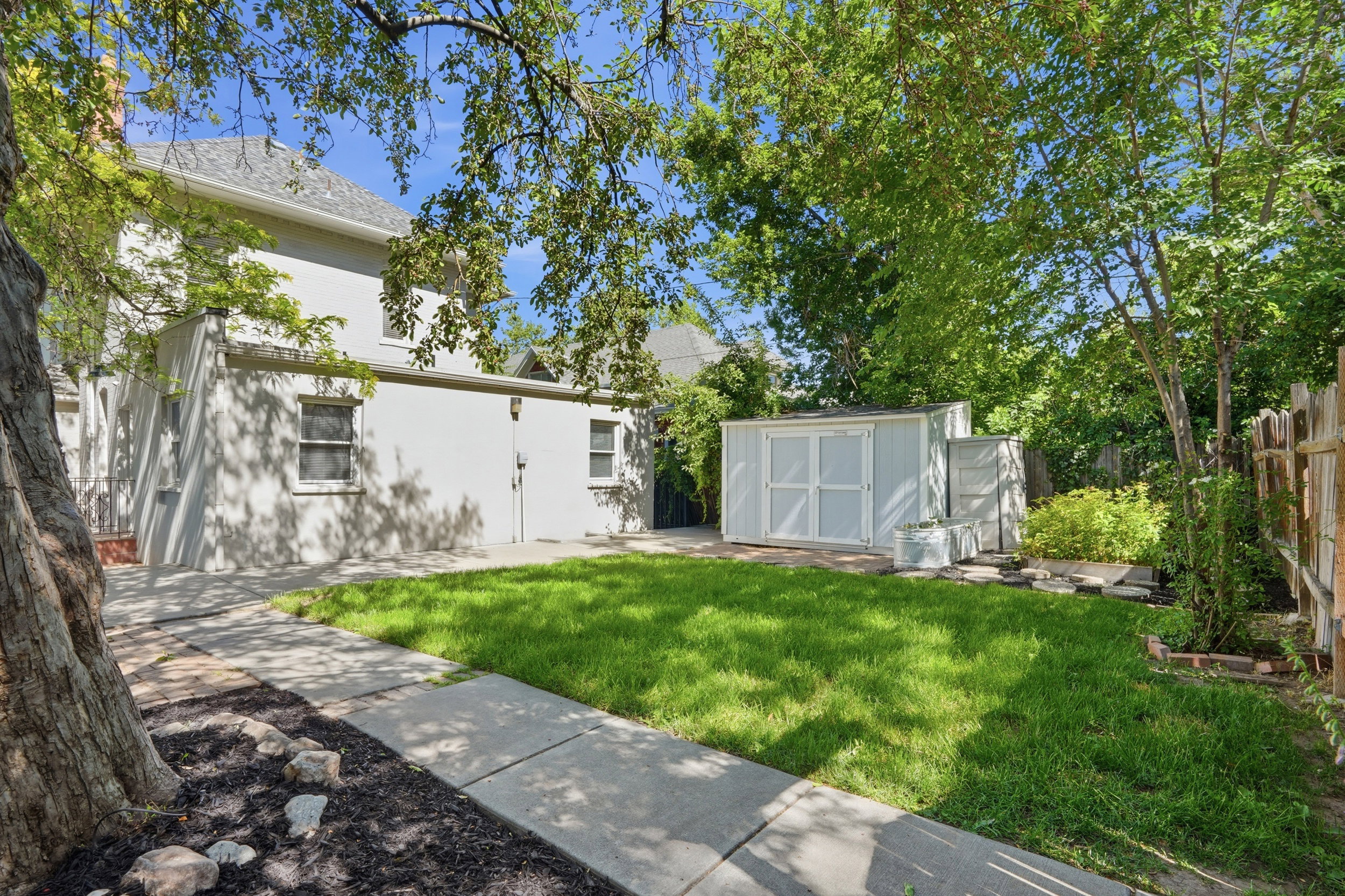 Expansive backyard featuring a green lawn, concrete pathway, mature trees, and a light-colored storage shed