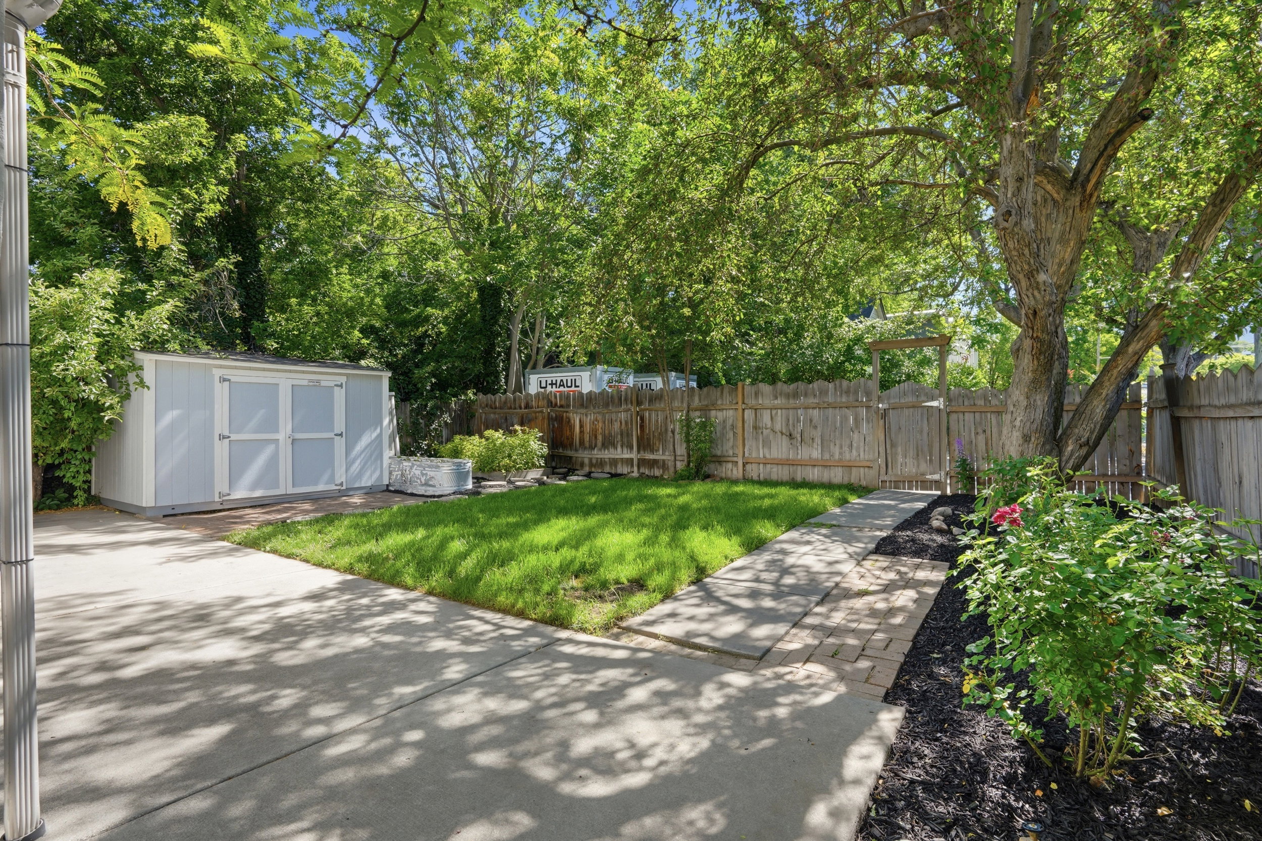 Spacious backyard featuring a light blue storage shed, a concrete patio, a stone paver pathway, mature trees, and a wooden privacy fence