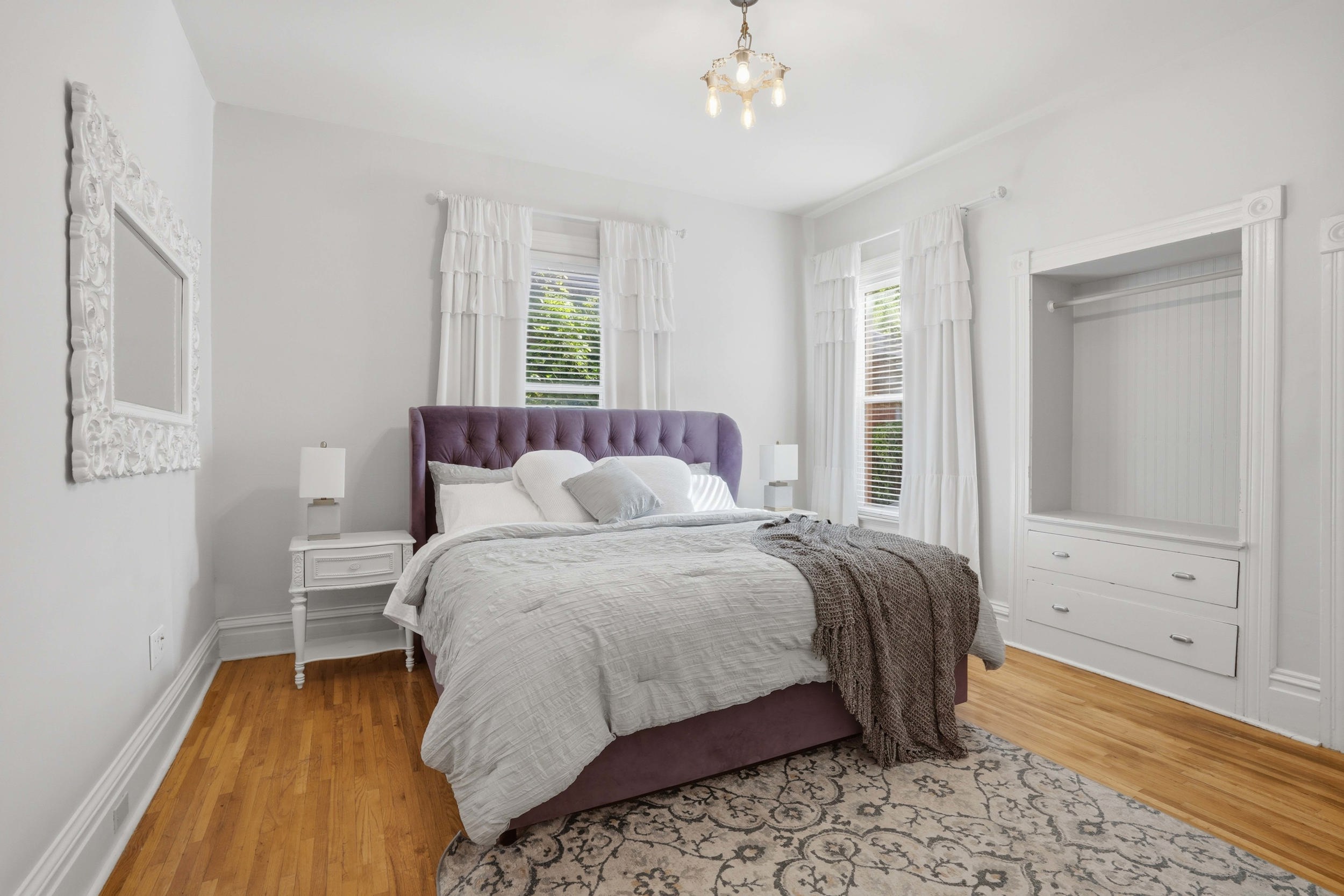 Bedroom featuring wood-finish flooring, two large windows with blinds, a built-in wardrobe with beadboard backing and two drawers, and a ceiling-mounted light fixture